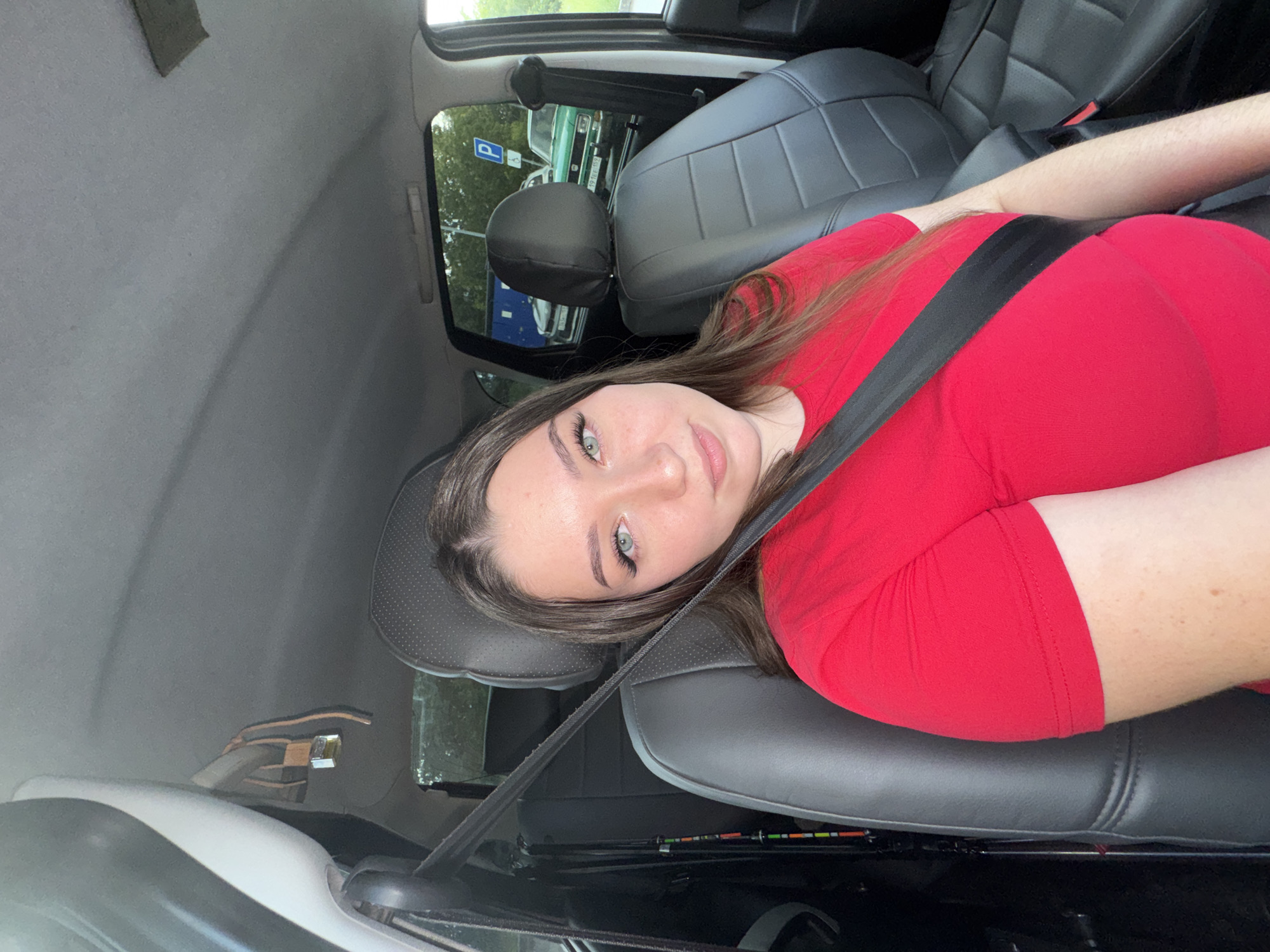 A young woman with long brown hair and light green eyes is sitting in the passenger seat of a car. She is wearing a bright red t-shirt and a black seatbelt. Her face is turned towards the camera, and she is looking directly at the viewer. The interior of the car is visible, including the black leather seats and the headrest. Outside the car window, trees and other vehicles can be seen, suggesting an outdoor location. The lighting indicates it is daytime. There is a small padlock hanging from the car's ceiling.