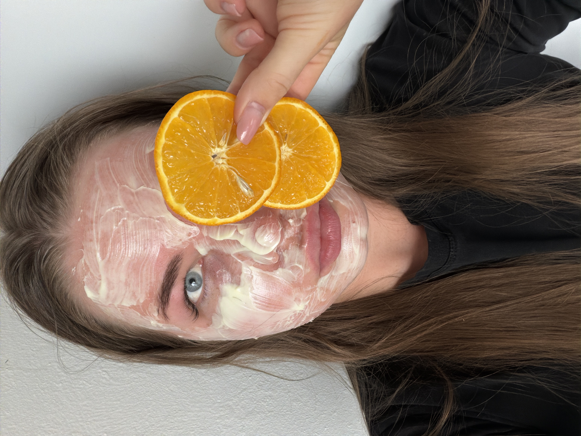 A young woman lies on a white surface, possibly a bed or examination table. Her face is covered in a thick, white cream, suggesting a facial mask. A manicured hand holds two orange slices over her forehead, obscuring part of the mask. The woman has light brown hair and blue eyes. She is wearing a black shirt. The environment is plain and lacks distinguishing features. There is no evidence to suggest the location is specifically in Chaikovsky, Russia.