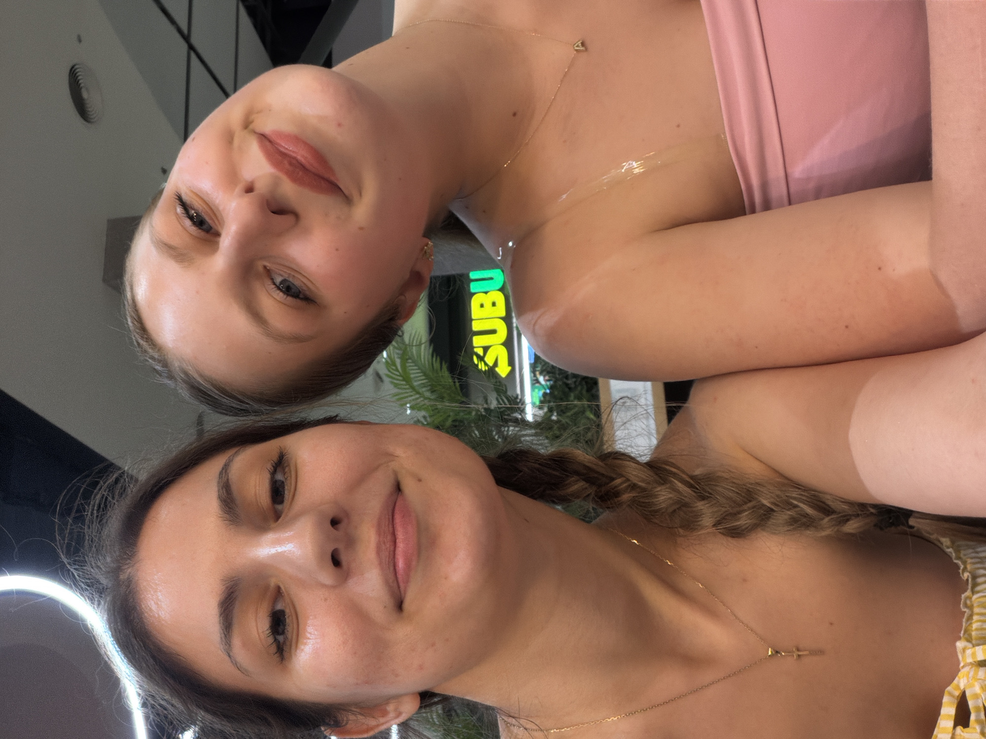 A close-up selfie captures two young women indoors. The image, oriented vertically, shows them from the chest up with subtle, friendly smiles.
The woman on the left (when viewed with correct orientation) has blonde hair and blue eyes, wearing a pink strapless top and a delicate gold necklace with a heart pendant. Her arm shows a distinct tan line. The woman on the right has dark brown hair styled in a braid over her shoulder and brown eyes, wearing a yellow and white striped top and a gold necklace with a small bar pendant. Her arm also displays a tan line.
In the background, a brightly lit neon green sign, partially visible as "SUBU" with an arrow, indicates a "SUBWAY" restaurant. Green foliage is also visible behind them. A circular light source, possibly a ring light, reflects in the eye of the dark-haired woman, suggesting artificial lighting for the photo. The setting appears to be a brightly lit indoor commercial space, such as a mall or food court, in Burgas, Bulgaria. Both women exhibit relaxed and pleasant expressions.