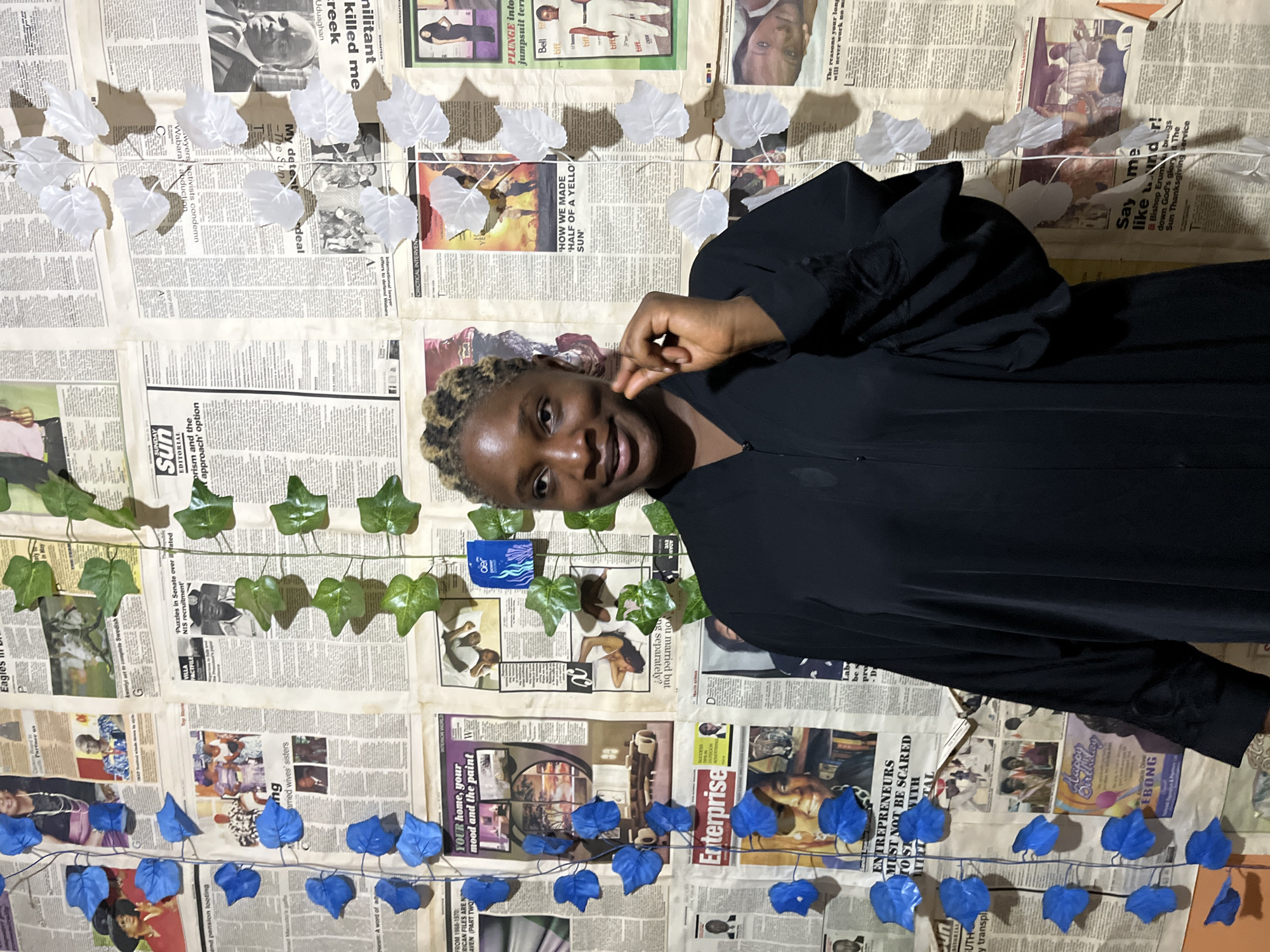 The image depicts a woman standing in front of a wall covered with newspaper clippings. The woman is wearing a black, flowing garment. She has her hair styled in braids and appears to be looking towards the camera, with her right hand near her face. The newspaper clippings on the wall are varied, featuring headlines, photographs, and advertisements. Decorative elements, including blue and white artificial leaves, are attached to the wall, interspersed with the newspapers. The overall impression is that of a creative or artistic backdrop within an indoor setting. The prompt mentions the location of Rafin Par, Nigeria, but this information cannot be visually confirmed from the image alone.