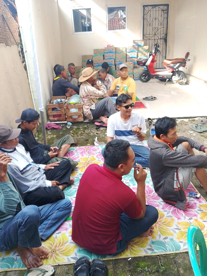 A group of men sit on a floral mat in a courtyard. Some men are seated against a wall lined with stacks of bottled water cases. A red and brown moped stands near a gated door. The men are dressed in casual clothing, and several wear hats or sunglasses. Some are engaged in conversation or looking at phones. The setting suggests a social gathering or community event.