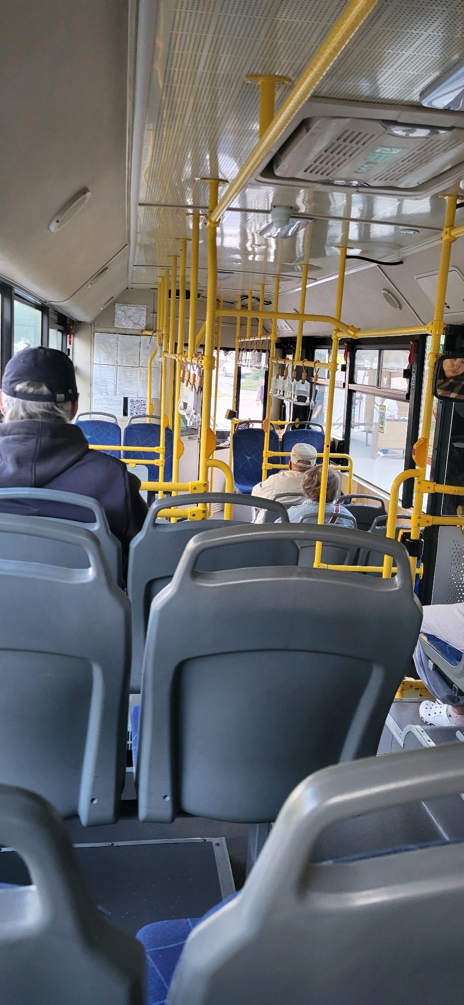 The image shows the interior of a public bus, likely in Oskemen, Kazakhstan, as indicated by the QR code and informational posters visible on the bus wall, which are commonly used for public transportation in Kazakhstan. The perspective is from a seated passenger looking towards the front of the bus. The seats are grey with blue fabric cushions. Yellow handrails and poles are prominent throughout the bus, designed for passengers to hold onto.
Several passengers are visible. In the foreground, the backs of two grey seats are shown. Further ahead, a passenger wearing a dark hoodie and a dark baseball cap is seated, facing forward. To the right of this passenger, another passenger wearing a light-colored cap and jacket is also seated. Near the back of the bus, another passenger's head and shoulders are visible. The windows of the bus are large, allowing natural light to enter, and some greenery can be seen outside. The ceiling of the bus has an air conditioning unit and lighting fixtures. The overall impression is a typical scene on a public bus during the daytime.