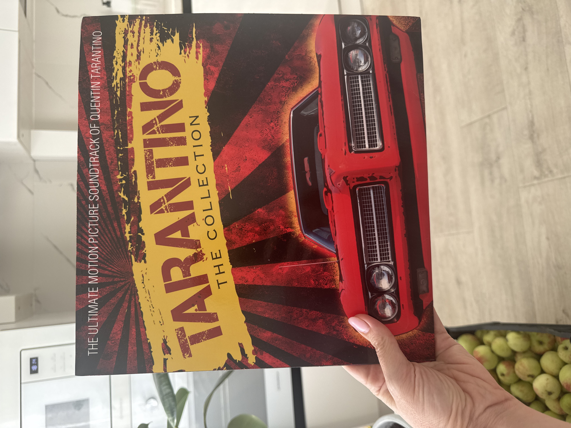 A person holds a vinyl record titled "TARANTINO THE COLLECTION" in front of a microwave oven. The record features a red vintage car against a sunburst background. The person's hand with pink-painted nails is visible at the bottom left of the record. In the lower left corner of the image, a black crate filled with green apples is partially visible. The background includes a white kitchen counter and cabinetry. The image does not provide any information about the city of Ufa, Russia.