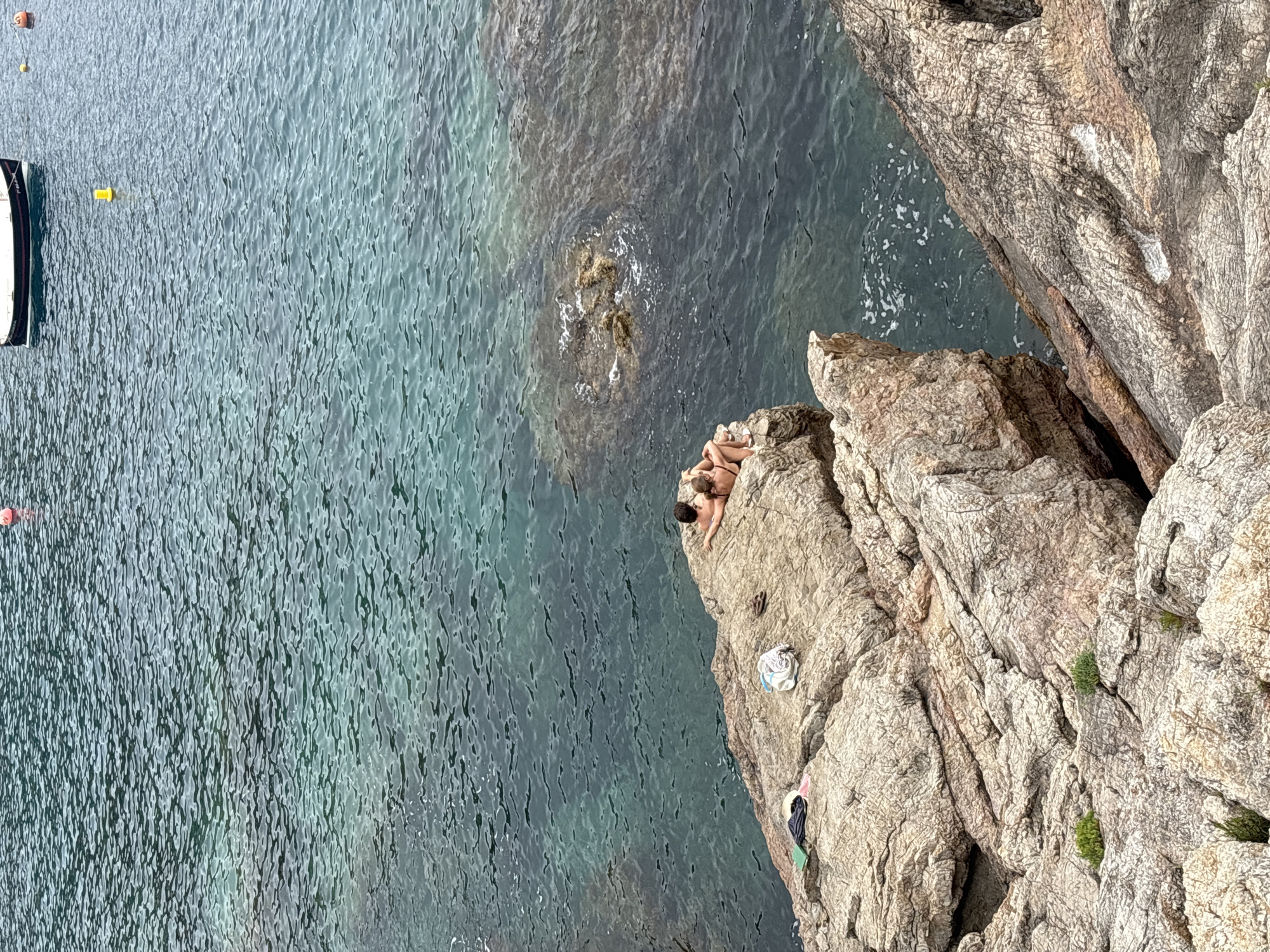 Two individuals are seated on a rocky outcrop overlooking the sea in Begur, Spain. The individuals appear to be a couple, one resting their head on the other's shoulder. They are dressed in swimwear. Scattered on the rocks are personal belongings, including a hat, a towel, and a pair of flip-flops. The sea is calm and clear, with visible ripples on the surface. A boat is visible in the distance, anchored in the water. The rocky terrain suggests a coastal environment, typical of Begur's coastline.