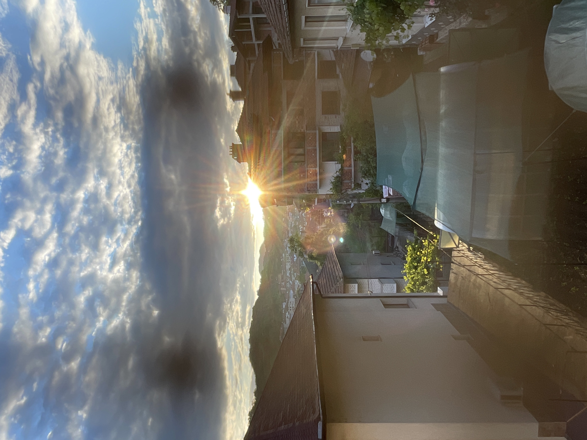 The image presents a detailed, elevated view of Zlatograd, Bulgaria, at sunset. The sky, occupying the upper third, features a mix of dark stratus clouds across the center and brighter altocumulus clouds above, with clear blue sky visible in the upper right. The setting sun, positioned centrally on the horizon line, emits prominent lens flares and bright, golden rays that extend into the cloudy sky and across the urban landscape below, casting long shadows.
In the midground, a dense cluster of multi-story residential buildings with traditional red-orange tiled roofs and light-colored facades dominates the scene. Several structures feature balconies, some adorned with greenery, and a visible satellite dish and an air conditioning unit are mounted on exterior walls. One building towards the upper right midground appears to be under construction or partially exposed, showing brickwork. In the background, verdant, undulating hills delineate the city's periphery.
The immediate foreground, seen from a high vantage point, includes the corner of a large, light-colored building with a brown tiled roof on the left. Below and to the right of this building, a concrete ramp or walkway with a dark metal railing descends. Below this, two prominent green shade structures or greenhouses are visible, indicating agricultural or gardening activity. No individuals are present in the frame. The overall lighting suggests a tranquil, late afternoon or early evening atmosphere.