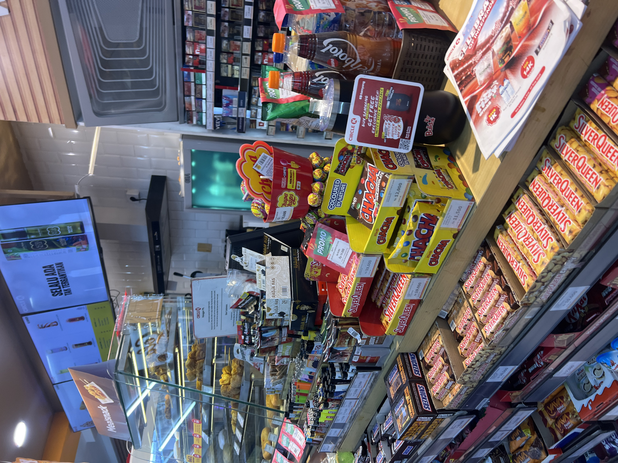 The image shows the interior of a Circle K convenience store in Denpasar, Indonesia. The store features a well-stocked display of snacks, candies, and drinks. Prominently displayed are various brands of chocolate bars, including SilverQueen and Snickers, as well as Chupa Chups lollipops. There are also bottles of drinks, including what appears to be a brand called "Ichbotol". In the background, shelves are filled with cigarettes and other packaged goods. Above the counter, digital screens display advertisements. The overall environment is brightly lit, typical of a convenience store setting.