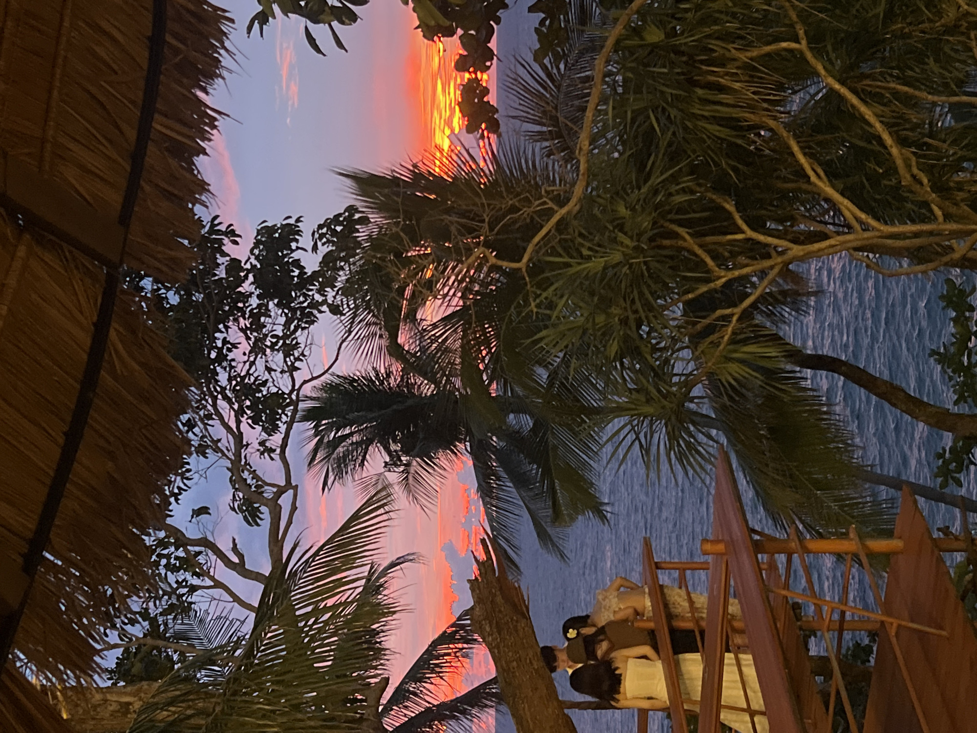 The image captures a vibrant sunset over the Andaman Sea, viewed from an elevated wooden platform in Karon, Thailand. In the foreground, a thatched roof structure partially obscures the sky, with the silhouette of palm trees framing the scene. On the platform, three individuals, two women and one man, are visible from behind, dressed in formal attire. They appear to be observing the sunset. The sea is a deep blue, with gentle waves, and the sky is a dramatic display of orange, pink, and purple hues. The overall impression is one of serene beauty and a romantic evening setting.