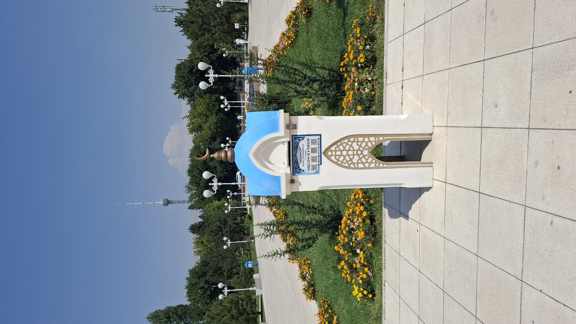 The image shows a small, white, mosque-like structure with a blue dome and a gold crescent moon on top, located in a park. The structure has an arched opening and a decorative facade. It is surrounded by green grass and a flower bed with yellow and orange flowers. In the background, there are trees, lampposts, and a tall tower in the distance. The sky is clear and blue. The image is taken from a slightly low angle, looking up at the structure and the background elements.