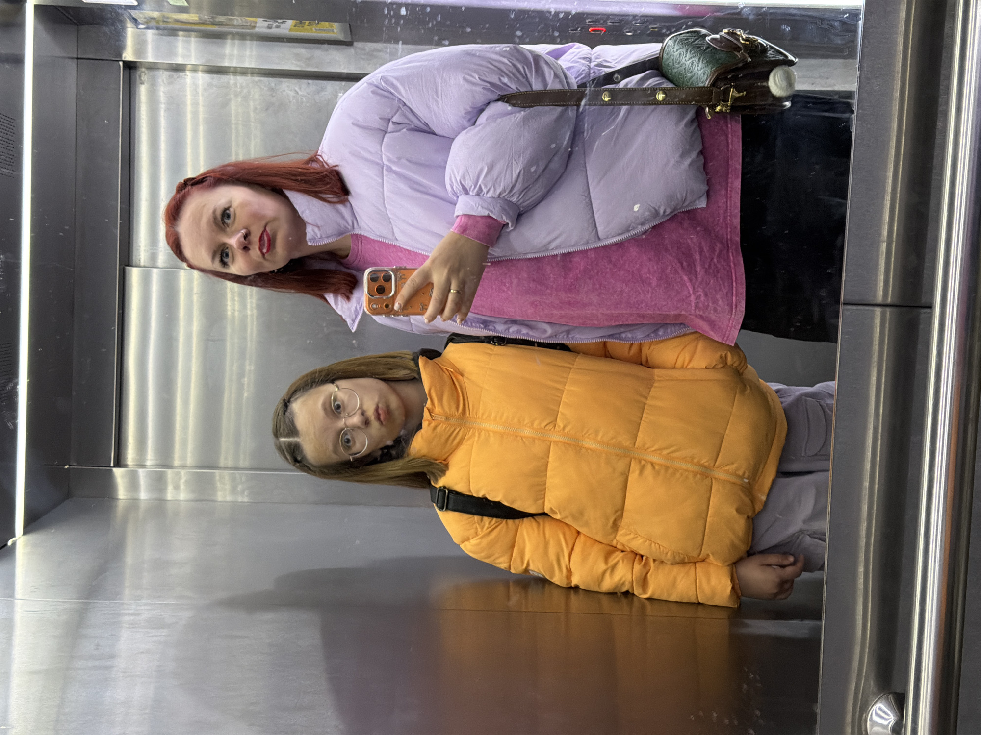 Two women are reflected in a silver elevator. One woman has red hair and is wearing a purple puffer jacket and a pink shirt, holding a phone. The other woman has blonde hair and is wearing glasses and an orange puffer jacket. The elevator walls are metallic. The location is Unknown, Unknown.
