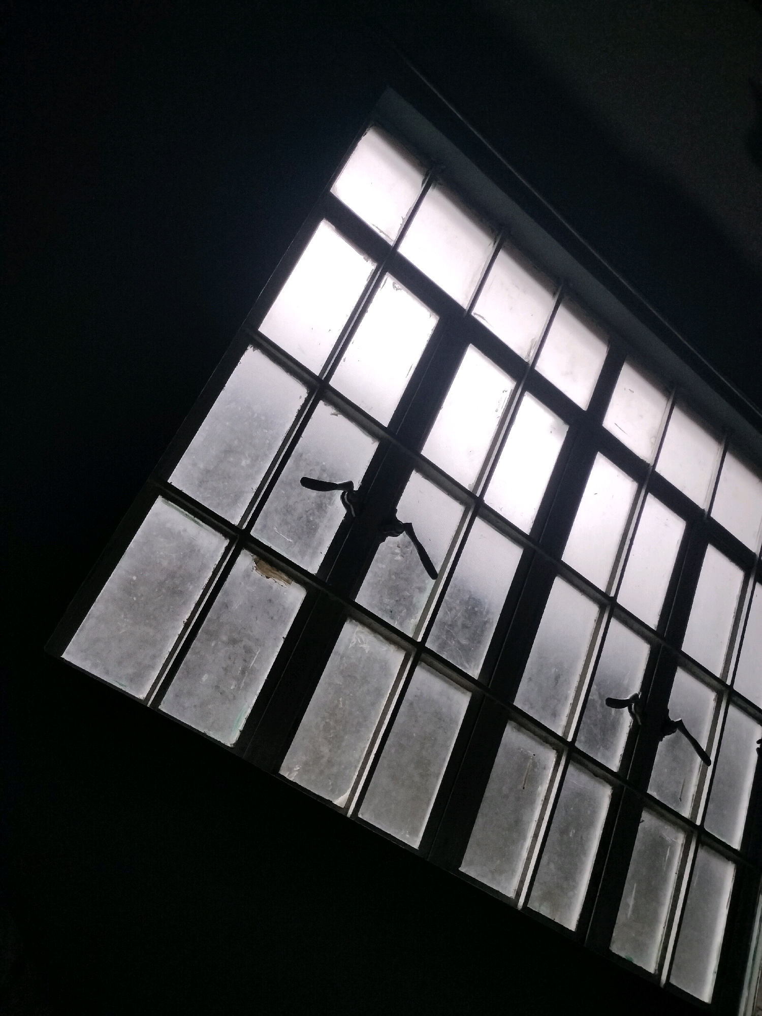 A tilted, wide-angle shot looks up at a large, grimy industrial window from below and to the side. The window, composed of many small rectangular panes divided by dark metal mullions, takes up most of the frame. The panes are covered in dust and streaks, obscuring the view of what is outside. Two black metal window latches are visible on the windowpanes. The window is set against a very dark, unlit background. The overall scene evokes a sense of neglect or abandonment. The location is identified as the city of Baler, Philippines.