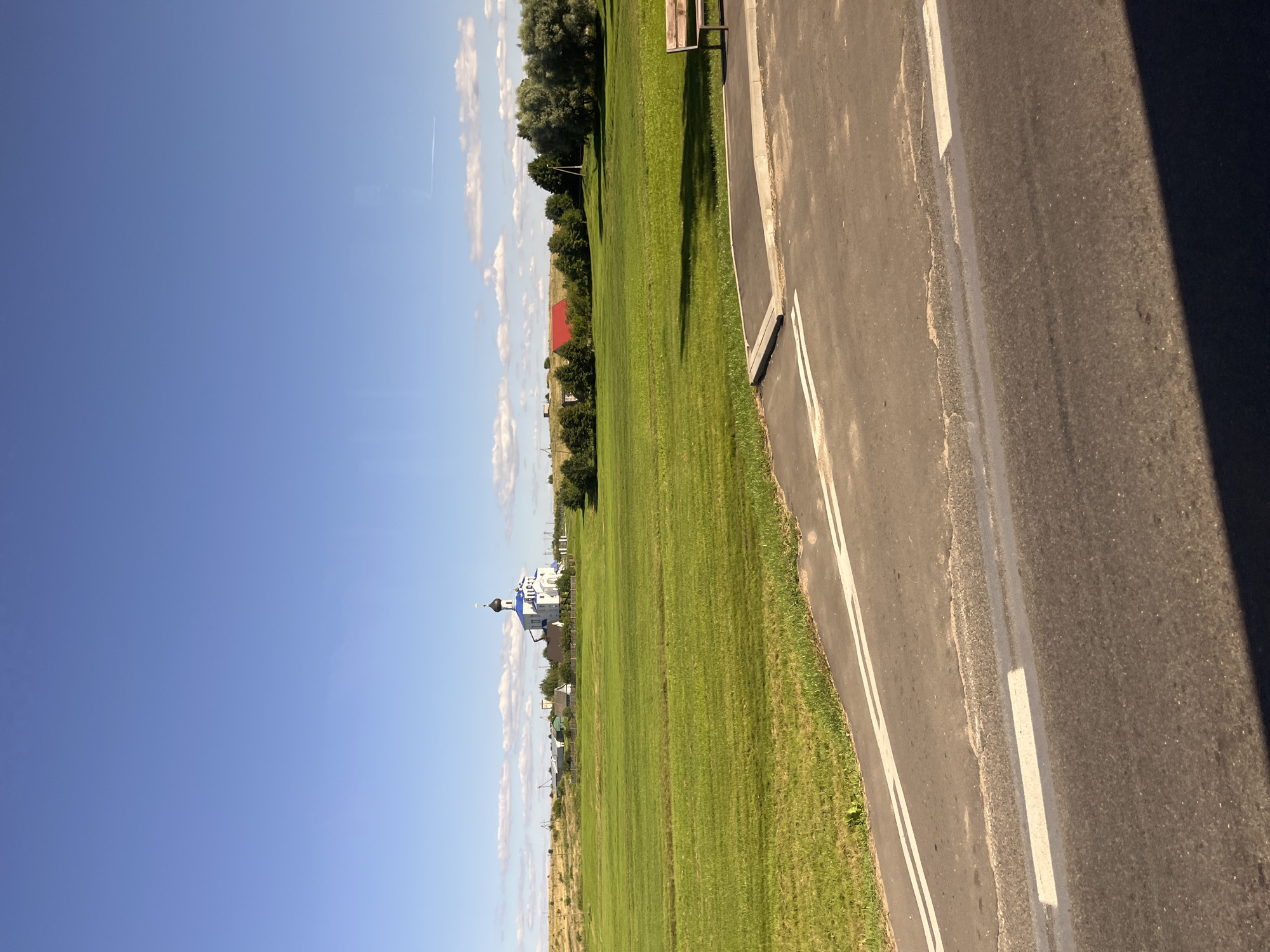 The image depicts a rural landscape under a clear blue sky with scattered white clouds. In the mid-ground, a white church with a blue dome and a black cupola stands prominently, surrounded by a cluster of buildings and a white fence. Adjacent to the church is a large, neatly mowed green field. To the right of the field, a red-roofed building is visible, along with a line of trees and a solitary lamppost. A wooden bench is positioned on a paved area at the edge of the field, partially shadowed by the foreground. The foreground consists of a paved surface with white double-line markings, suggesting a road or pathway. The scene appears to be located in or around the city of Myadzyel, Belarus.
