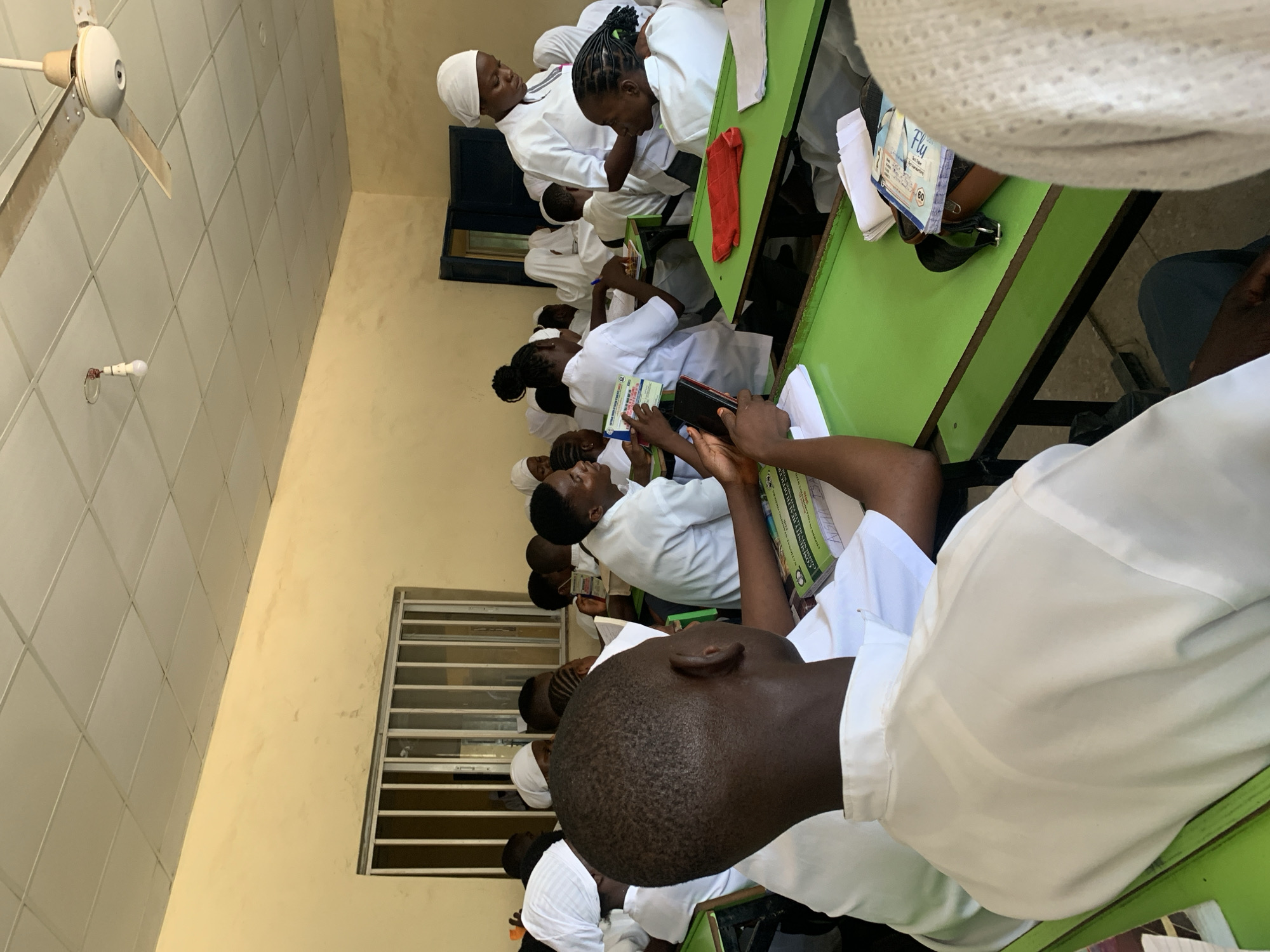 The image shows a classroom scene in Keffi, Nigeria. Several individuals are seated at green desks, primarily dressed in white uniforms. A few wear white head coverings. They appear to be engaged in reading or writing, as indicated by the presence of books and notebooks on their desks. One individual is looking at their cell phone. The classroom environment includes a window with bars, a ceiling fan, and tiled walls. The interior walls are painted in a light beige color.