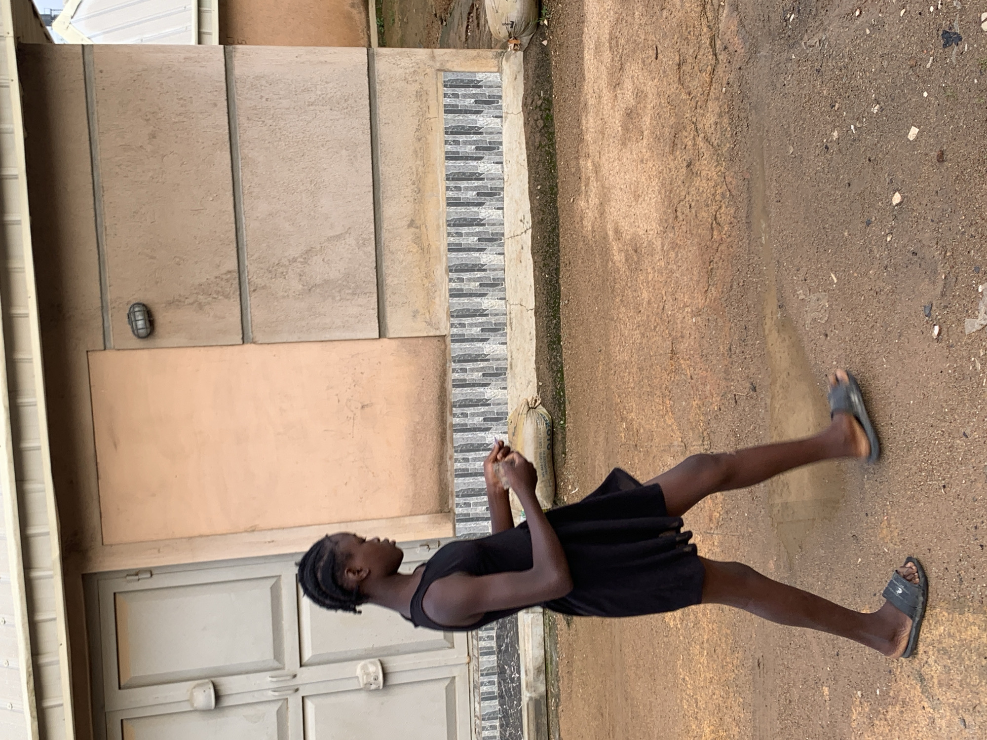 A young girl with braided hair, wearing a black sleeveless dress and dark sandals, walks from left to right across a dirt path in front of a light brown building in Jos, Nigeria. She is holding something in her hands, which are held in front of her. The building has a cream-colored overhang at the top and double doors on the left side. A strip of decorative tiles runs along the lower part of the building. The ground is a mixture of dirt and gravel, with some puddles of water. A large, lumpy, sand-colored sack lies on the ground to the right of the girl. The lighting suggests it is daytime, possibly overcast.