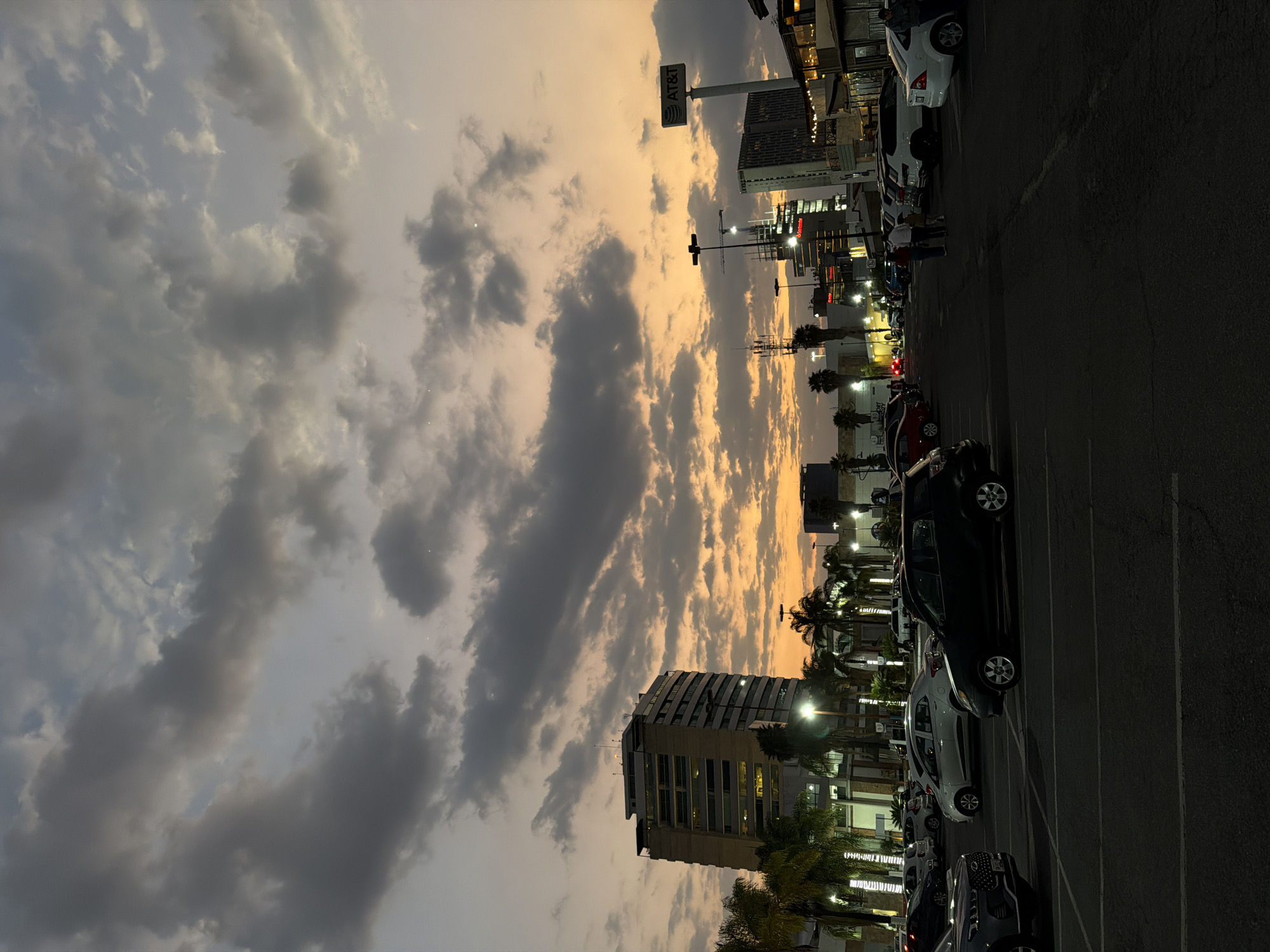 This image captures an urban landscape at dusk within San Bernardino Tlaxcalancingo, Mexico, focused on a large asphalt parking lot under a vivid sunset sky. The upper portion of the frame features a dramatic sky displaying an orange-yellow glow on the horizon, transitioning to light blue and dense gray clouds overhead.
The foreground consists of a dark asphalt surface with faded white painted parking lines. Numerous vehicles are parked across the midground: a dark gray SUV is partially visible on the far left, a black hatchback is positioned centrally, and a white sedan, a black SUV, and a white Ford Focus are among other cars on the right.
Several individuals are present in the midground-right, including a family unit consisting of an adult male, an adult female, and a child, walking away from the viewer. Other human figures are vaguely discernible near vehicles in the distance.
The background reveals a city environment. On the far left, a multi-story modern building, possibly an office or residential high-rise, features a beige and gray facade with numerous windows and rooftop structures. Palm trees, illuminated by artificial lights, are situated in front of this building. Toward the center-right, a prominent AT&T sign is displayed on a tall pole. Adjacent structures include commercial buildings with illuminated windows and outdoor seating areas. A building under construction, identifiable by visible scaffolding and a crane, is located further back on the right. Streetlights, power poles, and telecommunication towers are distributed throughout the background. Artificial lighting from buildings and streetlights is active, consistent with the low light conditions of sunset.