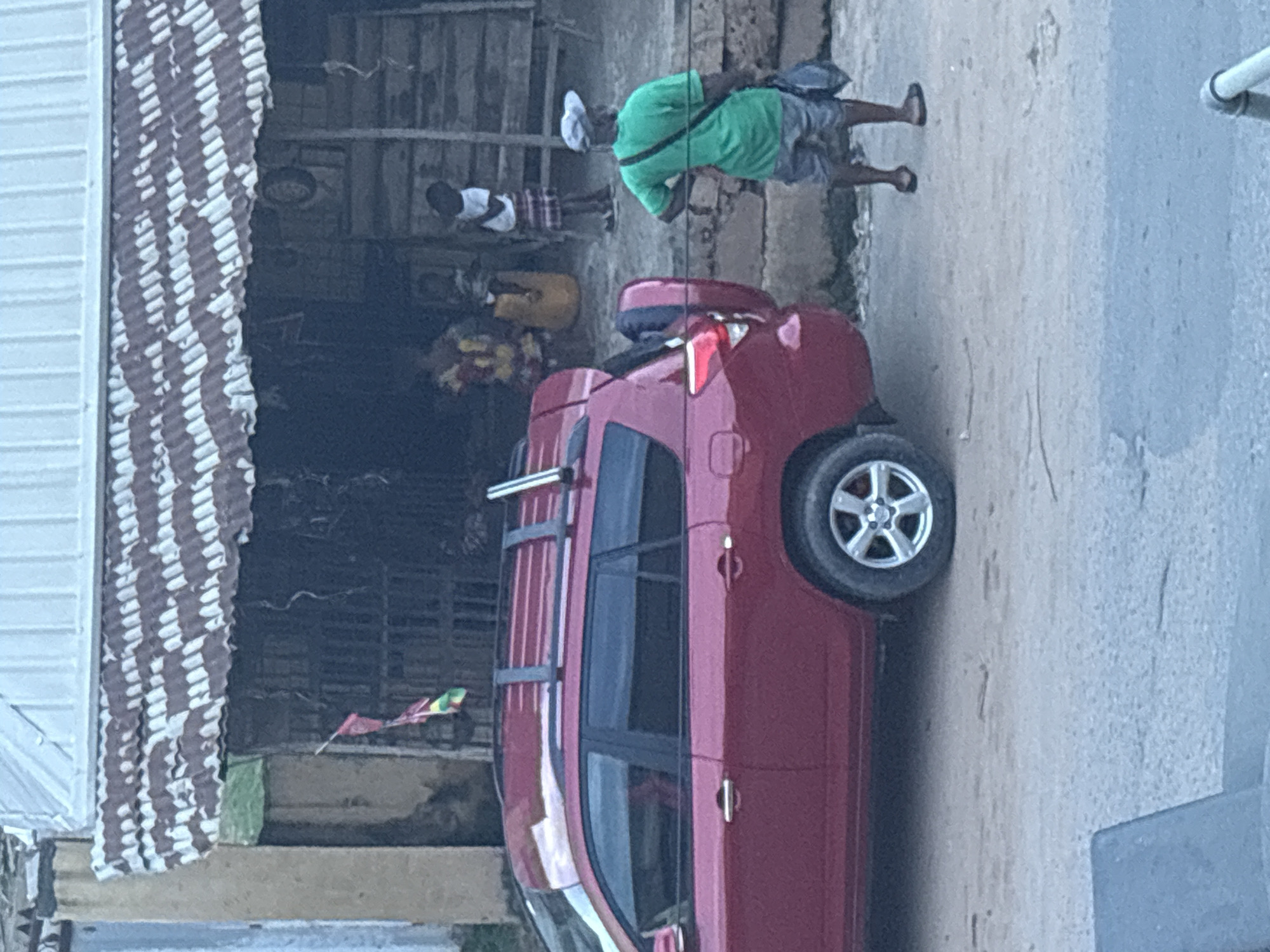 The image shows a street scene in Nkpor, Nigeria, with a red car parked on the side of the road. The car appears to be a modern hatchback with alloy wheels and a roof rack. In the background, there is a building with a corrugated metal roof and a open-fronted structure that might be a shop or market stall.
Several people are visible in the scene. One person in a green shirt and shorts is walking away from the camera, carrying a bag over their shoulder. Another person, wearing a white shirt and checkered shorts, is standing further back, partially obscured. There are also glimpses of other individuals and what appears to be some colorful decorations or merchandise in the background. A small, colorful flag or banner is attached to a pole in front of the building.
The lighting suggests it could be daytime, possibly late afternoon given the shadows. The scene seems to be an urban or semi-urban environment, likely a commercial area given the presence of the building and potential market stalls. The overall atmosphere appears to be casual and everyday. No visible text is present in the image.