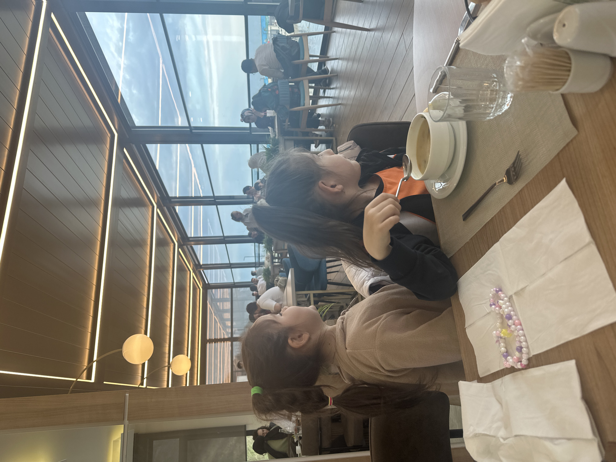 The image shows two young girls sitting at a table in a restaurant, possibly located in Arnavutköy, Turkey. One girl wears a black and orange jacket, while the other wears a tan sweater. On the table, there is a bowl of soup, silverware, a glass, a napkin, and a container of toothpicks. There is also a beaded bracelet. In the background, other people are seated at tables, and large windows overlook a body of water, presumably the Bosphorus. The restaurant has a modern design with lights along the walls.