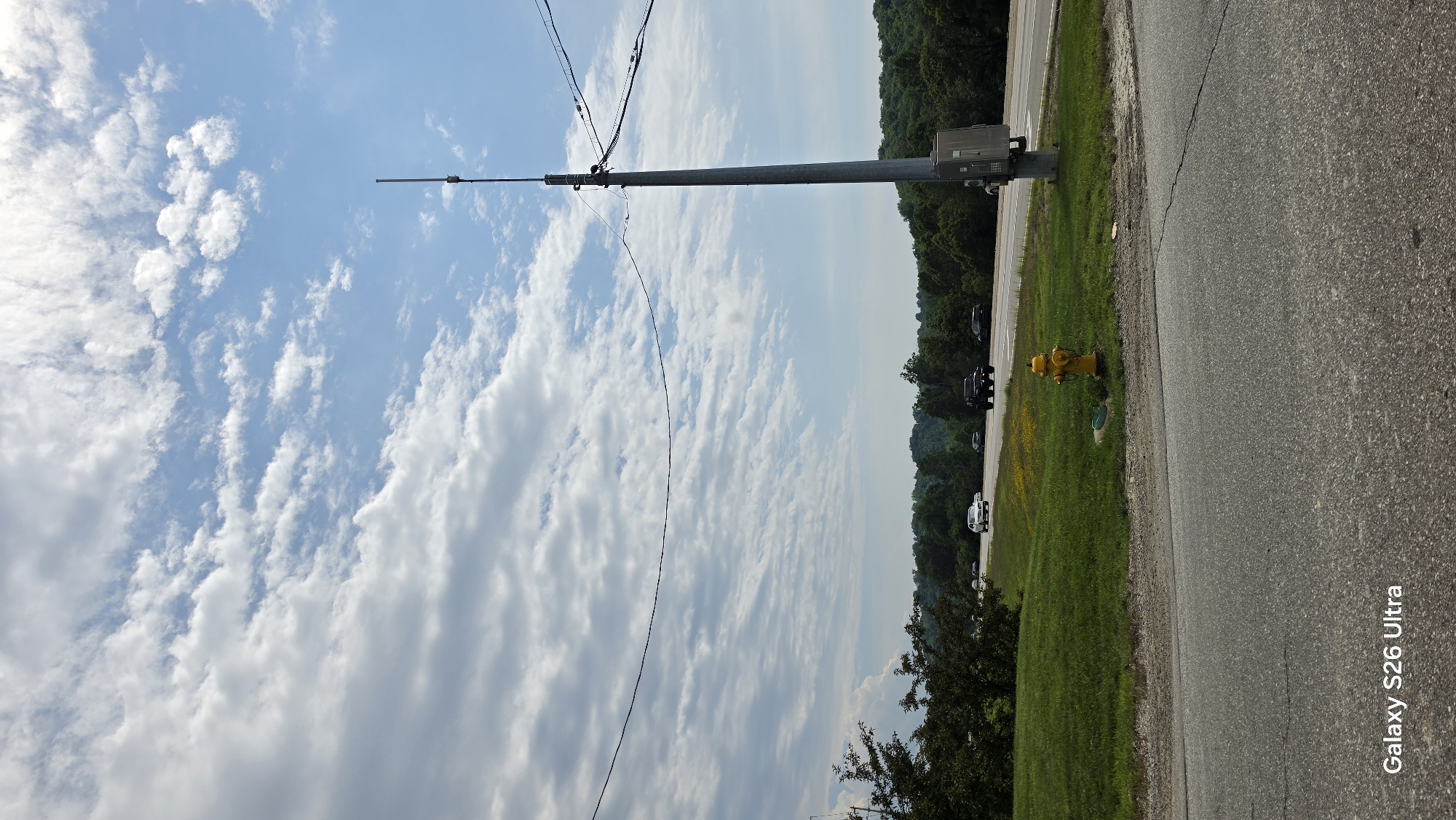 This media file depicts an outdoor scene with a clear sky filled with fluffy white clouds. A tall, grey pole with wires extends diagonally upwards from the left side of the frame towards the top right. Attached to the pole is a rectangular grey box.
To the right of the pole, a vibrant yellow fire hydrant stands out against a grassy area. Beyond the grass, a paved road runs parallel to the scene. Several dark-colored vehicles are parked or driving along the side of the road, with a white car further down the road. A dense line of green trees forms the background, suggesting a rural or semi-rural setting. The sun appears to be high in the sky, indicating it's likely daytime. The overall atmosphere is calm and ordinary, with no discernible emotional tone.
The visible text at the bottom right corner reads "Galaxy S26 Ultra," indicating the device used to capture the image. No people are clearly visible in the frame. The location context provided is Covington, United States of America.