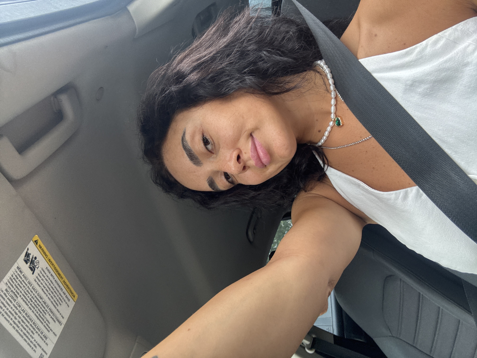A woman in a white top is taking a selfie in the passenger seat of a car, wearing a seatbelt. She has dark, wavy hair and is wearing a pearl necklace with a green pendant. The interior of the car, including the dashboard and a warning label on the sun visor, is visible. The background suggests an outdoor environment. The image was taken in Tbilisi, Georgia.