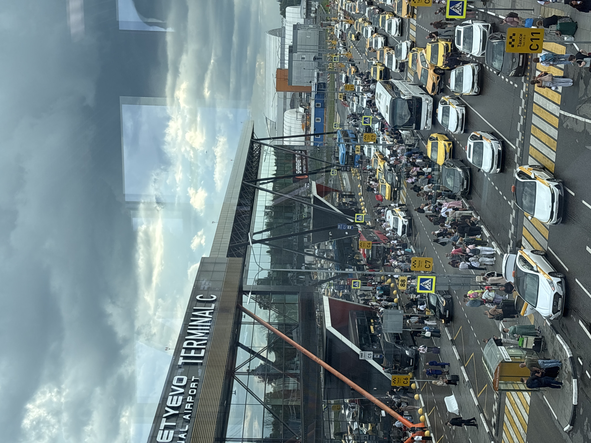 This media file captures a busy scene outside Sheremetyevo Terminal C, an international airport in Khimki, Russia. The image is taken from an elevated perspective, looking down at a long line of yellow and white taxis, along with other vehicles, queuing on the roadway. A crowd of people, many with luggage, are gathered on the sidewalk and near the taxis, suggesting the arrival or departure of travelers.
The time of day appears to be daytime, with a cloudy sky overhead, indicating overcast weather. The architecture of Terminal C is visible on the left side of the image, featuring a modern design with a glass facade. Road signs with text such as "Такси" (Taxi) and numbers like "C11", "C7", "C6", and "88" are prominent, indicating taxi zones and possibly departure gates. The overall atmosphere is one of bustling activity and movement associated with air travel.