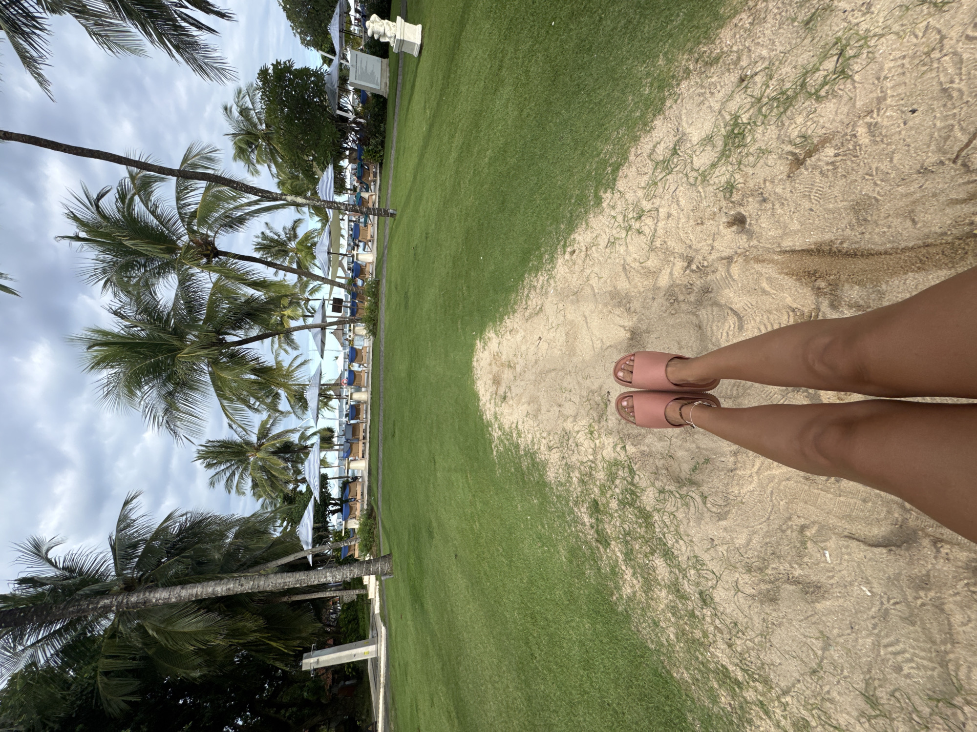 The image shows a person's legs in pink sandals standing on a sandy area next to a grassy lawn. Palm trees and beach umbrellas are visible in the background, suggesting a tropical beach resort setting. The location is likely in the city of Kuta Selatan, Indonesia, as stated in the prompt. The scene appears to be outdoors on a cloudy day.