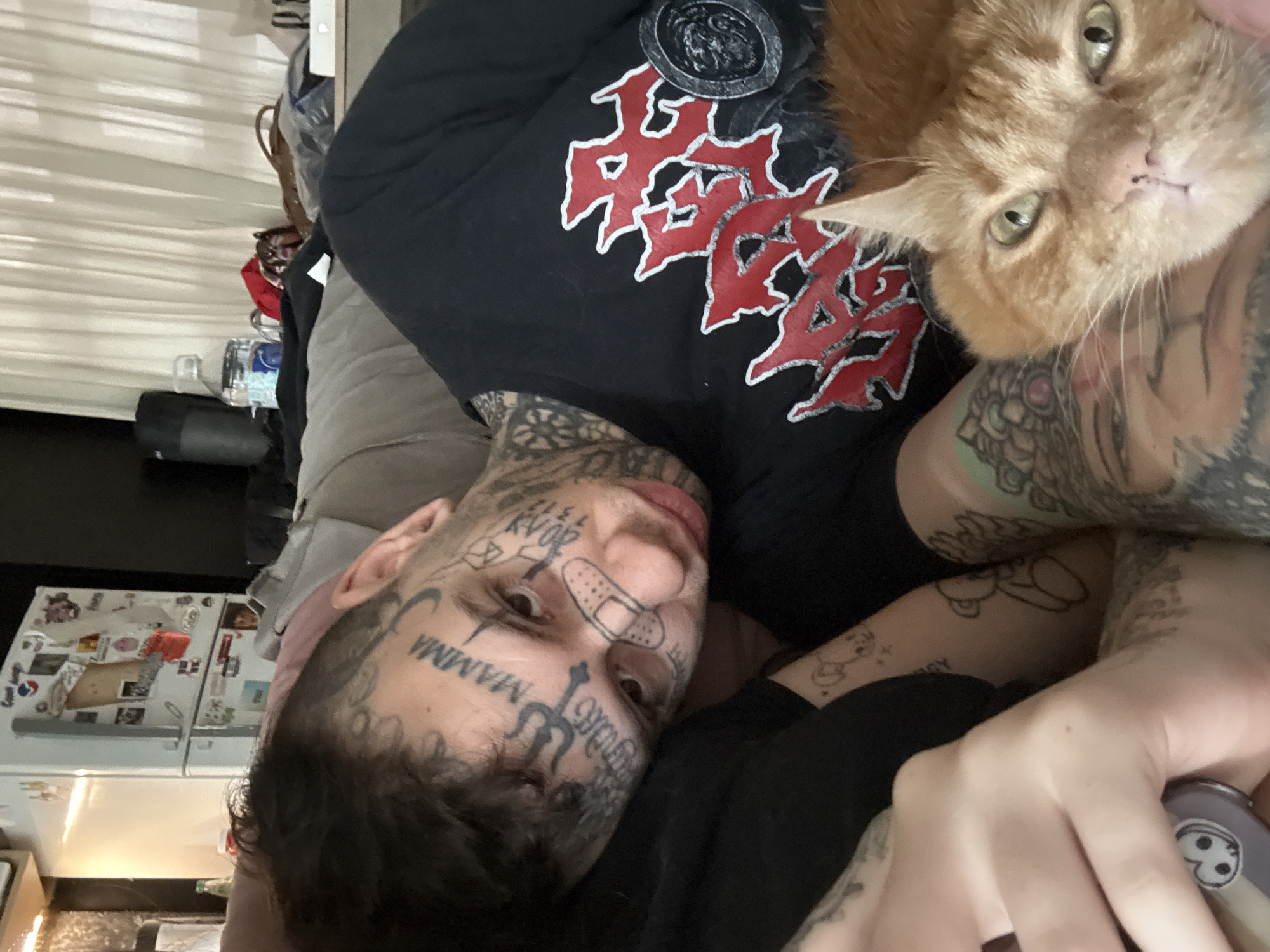 The image depicts an indoor scene in a domestic setting, located in Buenos Aires, Argentina. The primary subject is a male individual, shown from the chest up, reclining, with his head tilted towards the viewer. He has extensive facial tattoos, including the word "MAMMA" prominently on his forehead, a trident symbol, and other script and symbols, along with a drawn "band-aid" tattoo over his nose. His hair is dark and curly. He wears a black short-sleeved t-shirt featuring a large red and grey graphic, possibly text or a symbol, outlined in white. Visible on his arms are numerous tattoos, including abstract designs, a teddy bear, and other figures. His right hand, visible in the lower left, holds an object that is partially obscured, possibly a phone or remote, with a small cartoon-like sticker.
An orange tabby cat with green eyes rests directly in front of and below the individual's chest/shoulder area, looking towards the viewer.
The background reveals parts of a cluttered room. To the left, a white refrigerator is covered with numerous magnets, notes, and photos. A partial view of a light source is visible behind it. To the right, a large plastic water bottle, a black cylindrical object (possibly a speaker), and various items including bags and clothing are scattered on a surface, against a backdrop of light-colored, vertically-pleated curtains or blinds. The overall lighting suggests an interior daytime setting.