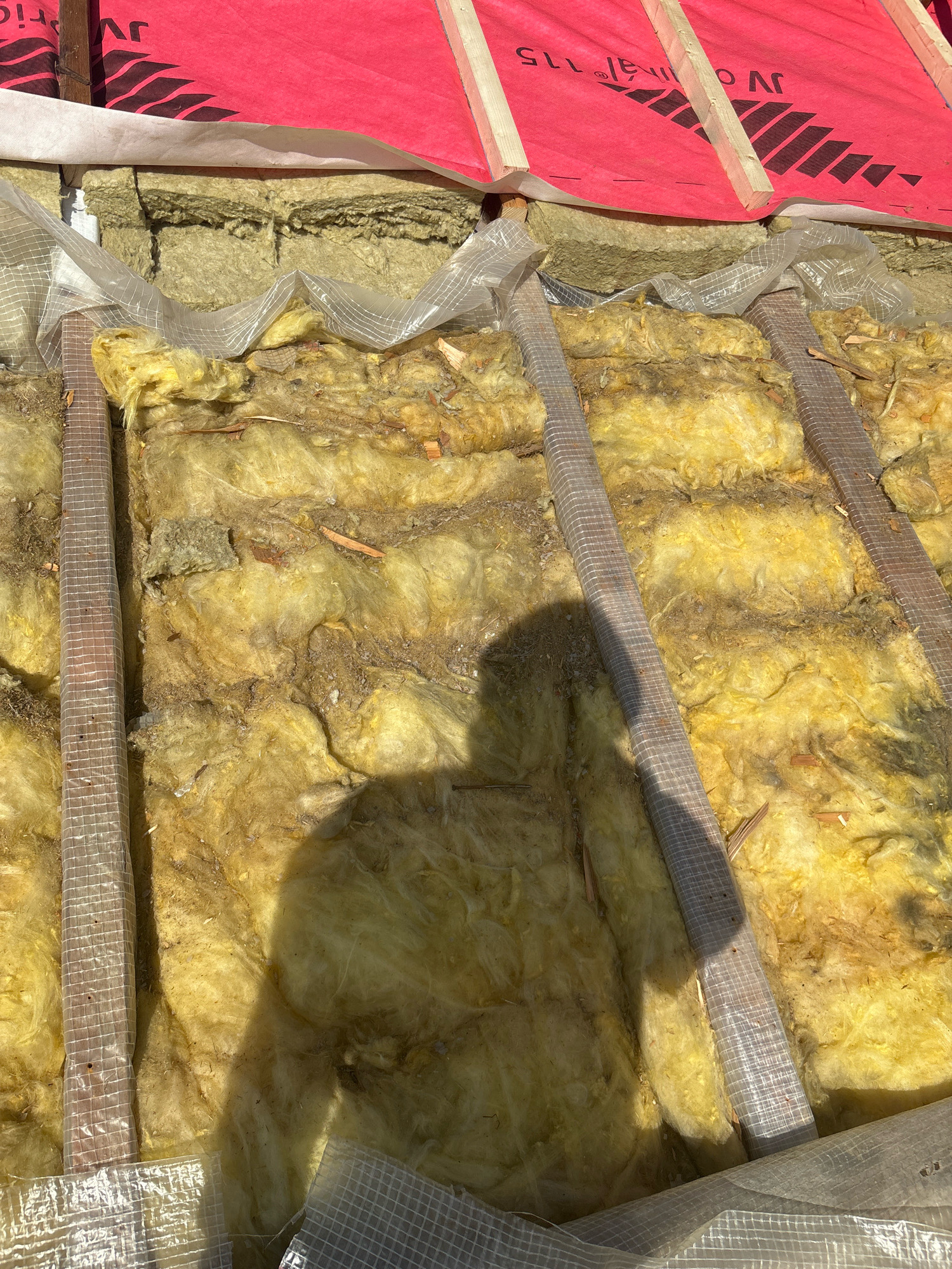 An exterior, medium-shot view of unfinished roof insulation in Zhavoronki, Russia. The insulation, composed of yellow fiberglass batting, is visible between wooden beams. A red building membrane with black lettering is stretched across the top of the frame, above the insulation. The scene appears to be a construction site, possibly a residential building. A shadow of a person is cast across the insulation.
