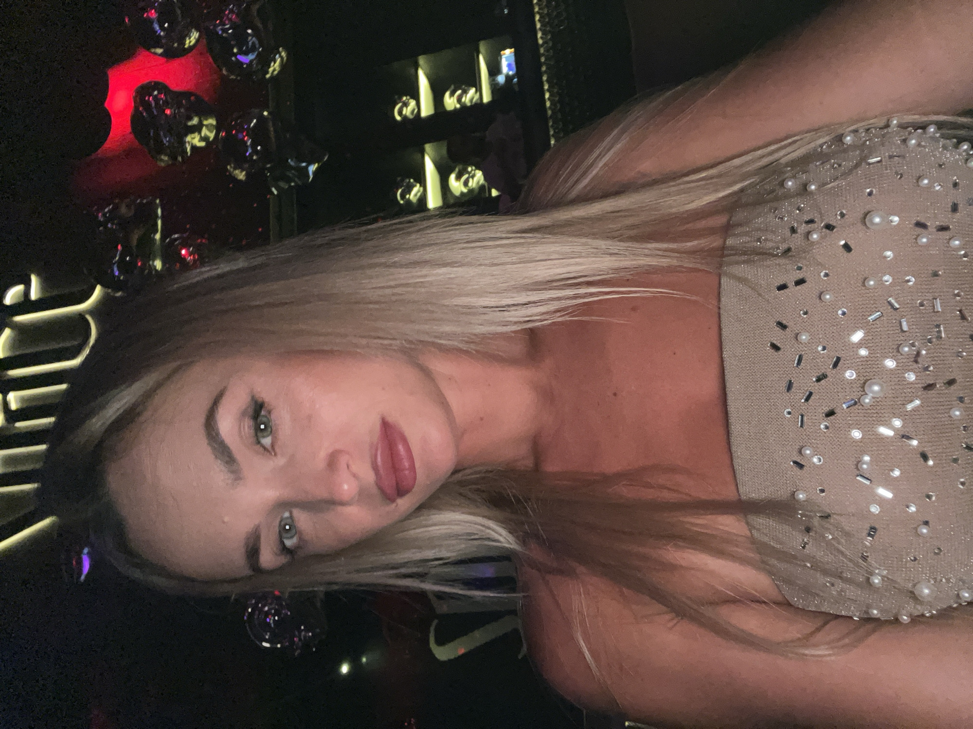 A woman with long blonde hair and fair skin is shown in a close-up portrait. She is wearing a strapless, embellished top. Her eyes are green, and she has full lips. The background is dark and appears to be an indoor setting, possibly a restaurant or club, with some decorative elements including lights and what look like metallic spheres. The lighting suggests it is nighttime. The image is a selfie.