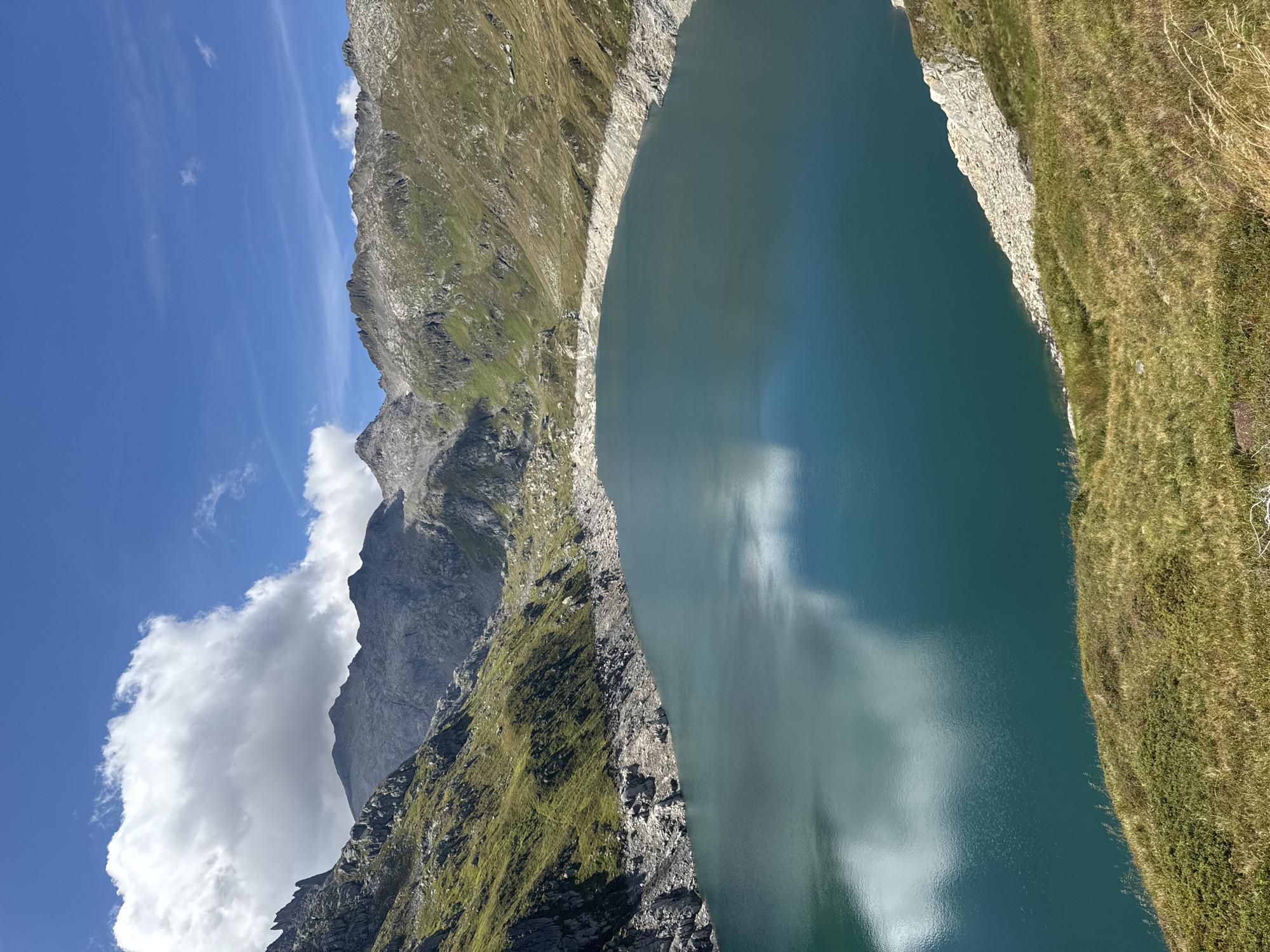 This is a landscape photograph of a reservoir lake nestled within a mountainous region. The lake is a deep, vibrant blue-green, with reflections of clouds and sky visible on its surface. The surrounding mountains are rugged and steep, with patches of green grass and rocky outcrops. The sky is bright blue with scattered white clouds, suggesting a sunny day. The foreground features a grassy hillside, adding a touch of natural texture to the scene. The location is the artificial reservoir of the Ritom dam, located in the Lepontine Alps, above Airolo in the canton of Ticino, Switzerland. The image captures a serene and majestic natural environment.