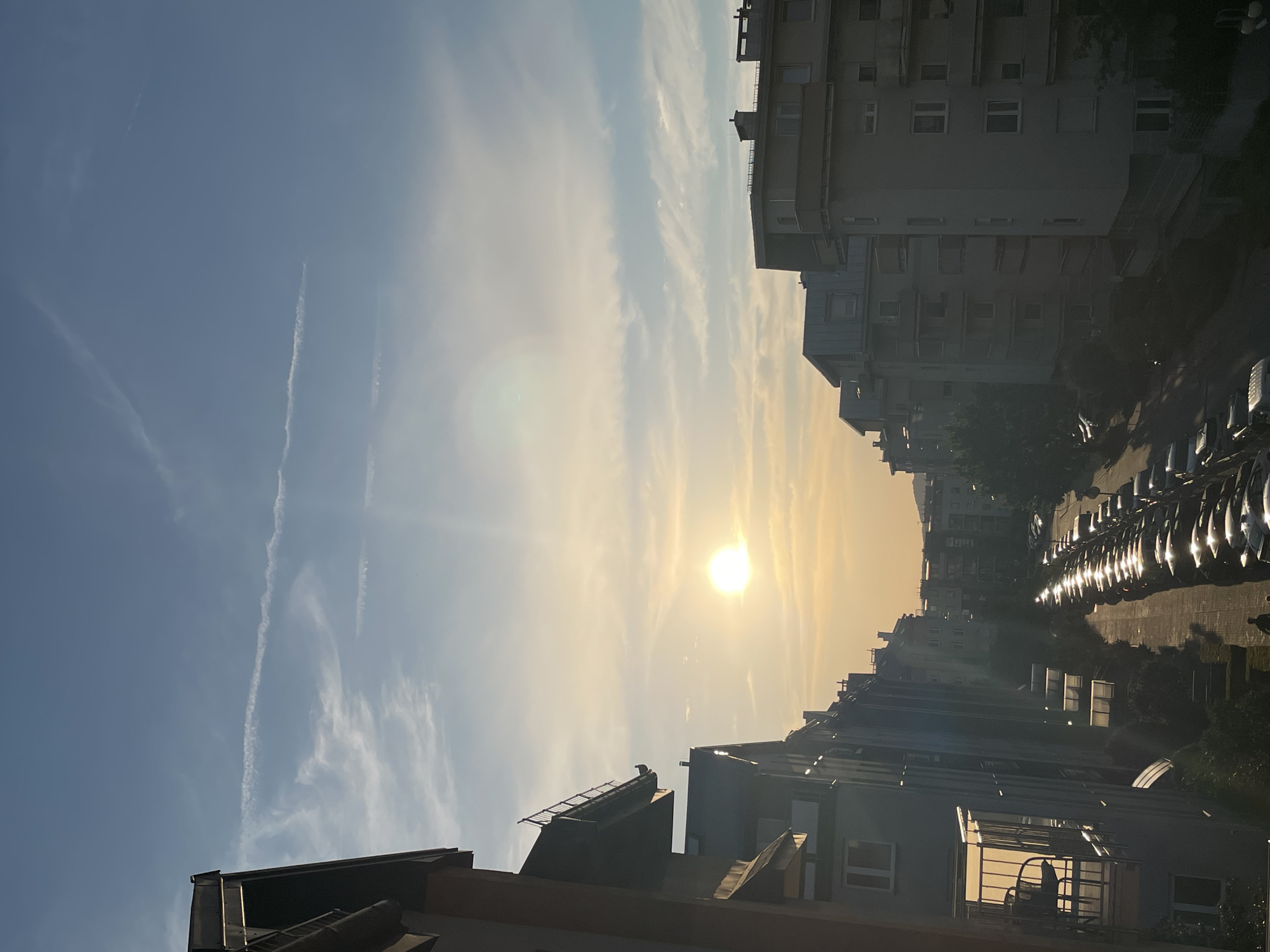 This media file captures a view of an urban street at sunset. The sun, a bright, diffused orb, is setting in the background, casting a warm, golden light across the sky. Wispy clouds streak across the clear blue sky, with faint contrails from airplanes visible.
The foreground and midground are dominated by buildings, appearing to be apartment blocks with multiple windows. Some of the buildings are silhouetted against the bright sky, while others are more clearly illuminated by the setting sun. A row of parked cars lines the street, their reflective surfaces glinting in the sunlight. The scene suggests a residential area within a city.
The time of day is clearly sunset, indicated by the low angle of the sun and the warm, golden hues in the sky and on the buildings. The weather appears clear with some cloud cover. There are no discernible people or specific activities taking place, lending the scene a quiet, tranquil atmosphere. The overall impression is one of a peaceful urban evening. No text is visible in the image.