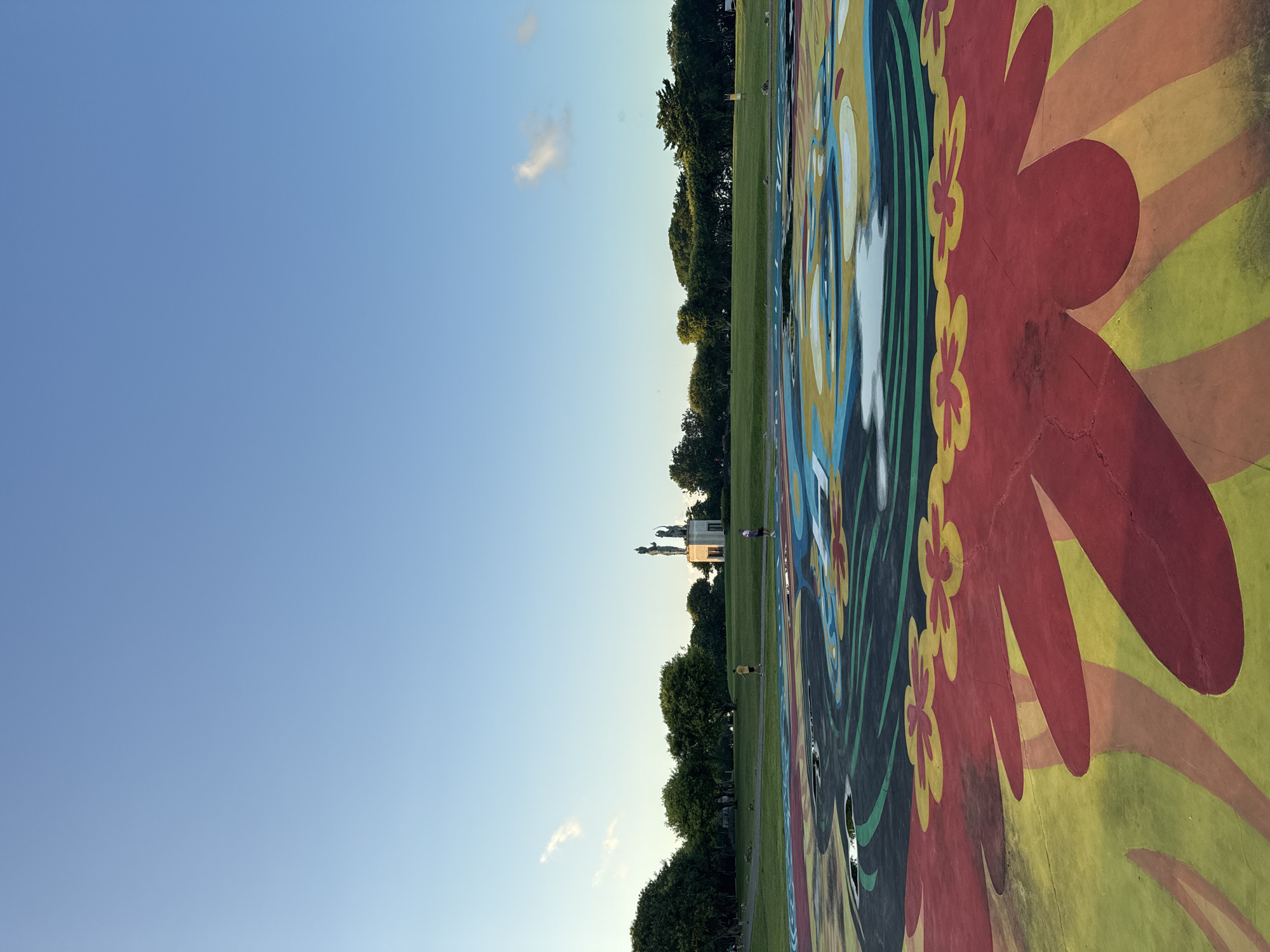 The image depicts a large painted mural on a ground surface, featuring floral and abstract designs in red, yellow, green, blue and black. A line of trees and a grassy field stretch horizontally across the center of the frame. The sky is a clear blue, with a small cloud. In the distance, there is a building with statues atop it. This mural is located in Garuda Wisnu Kencana Cultural Park in Kuta Selatan, Indonesia. There are two people walking near the building in the background.