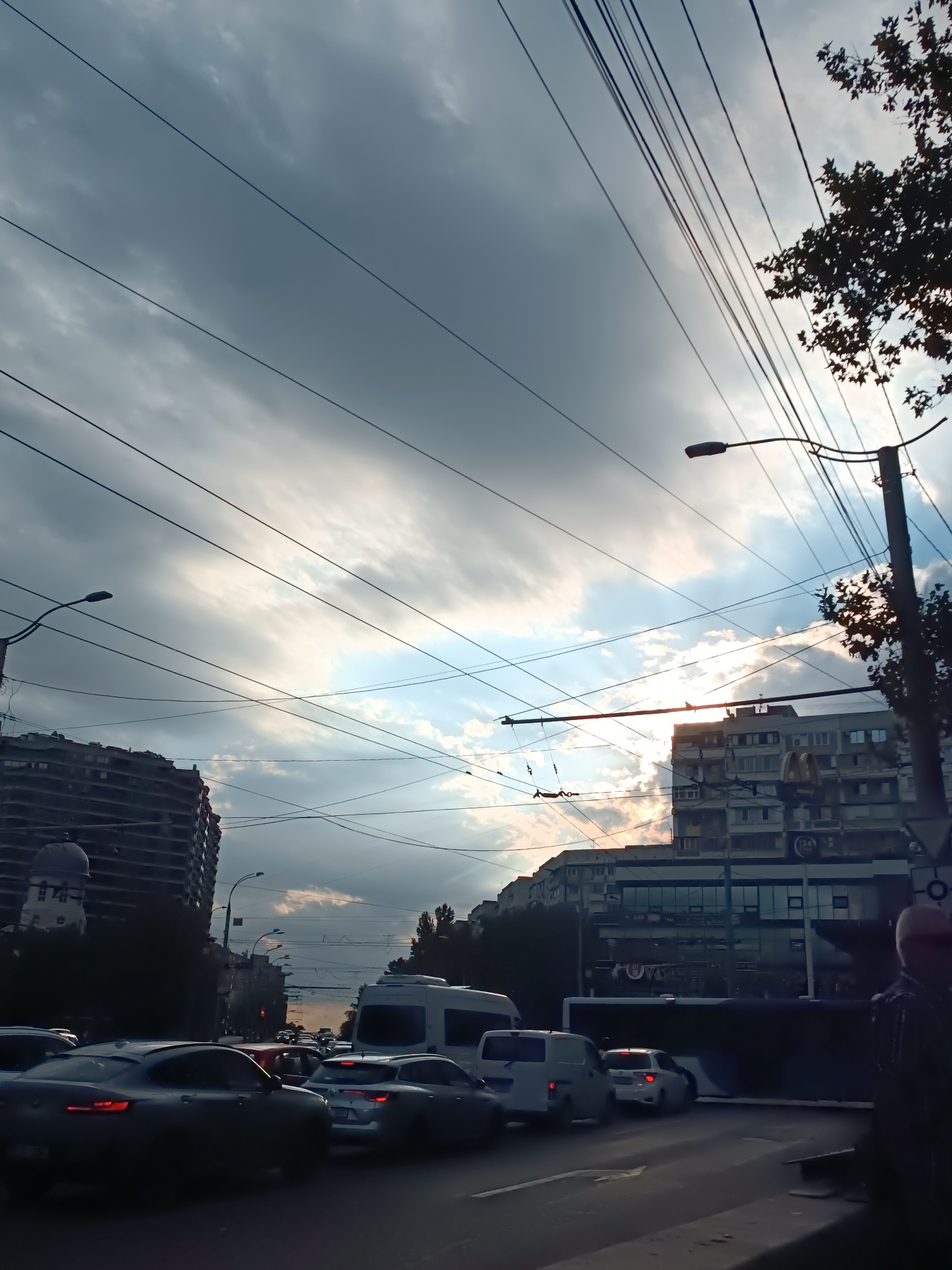 The image depicts a street scene in Chișinău, Moldova, with a focus on a traffic jam under a cloudy sky. Multiple vehicles, including cars, vans, and possibly a bus, are visible on the road, suggesting congestion. Overhead, a complex network of power lines and utility wires crisscrosses the frame, a common sight in urban environments. Buildings line the street, some appearing to be residential or commercial, with architectural styles that could be characteristic of Eastern European cities. The lighting suggests it is either early morning or late afternoon, with sunlight breaking through the clouds in some areas, creating a dramatic sky. The overall impression is of a typical day in a busy city, with the infrastructure of urban life and the flow of traffic dominating the visual.