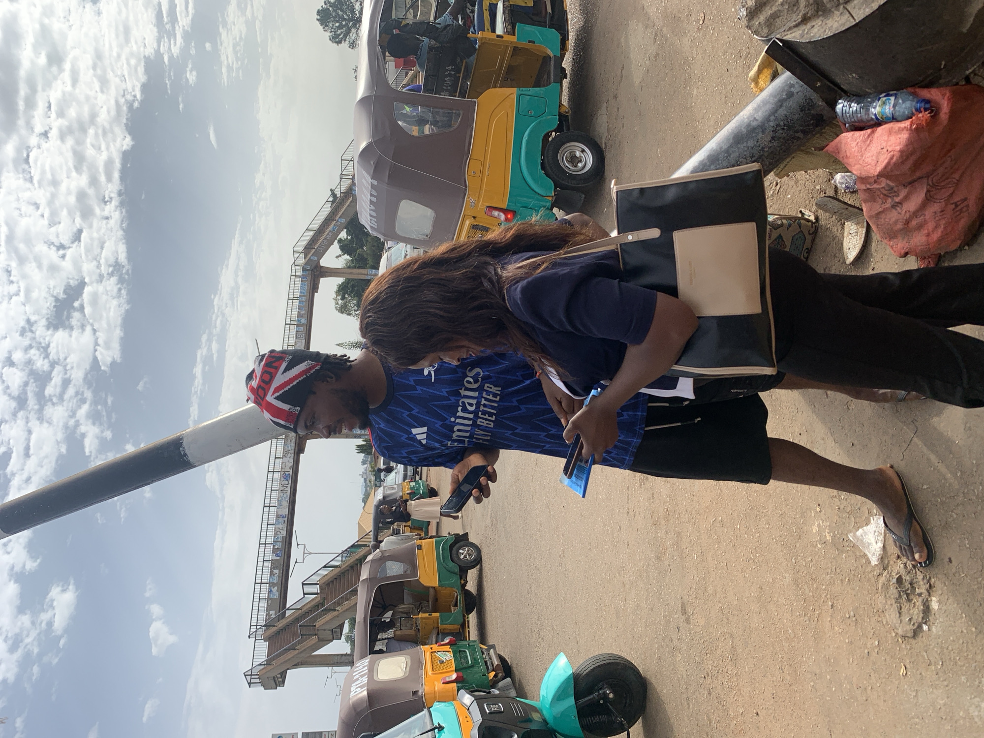 The image depicts an outdoor scene in Gura Top, Nigeria, with a focus on two individuals standing amidst a busy area with several motorized tricycles, commonly known as 'Keke Napeps'. The sky is partly cloudy.
In the foreground, a man wearing a blue and white patterned t-shirt and a beanie hat with a Union Jack design is holding a smartphone in his right hand. He is looking towards a woman who is standing to his left. The woman is wearing a dark blue top and dark trousers and is holding a blue item in her hands, possibly a phone or a small booklet. She is looking down at the item.
Behind them, several yellow and green Keke Napeps are visible, some parked and others appearing to be in operation. One Keke Napep has passengers inside. In the background, a pedestrian bridge crosses over what appears to be a road. A large, dark, cylindrical object is positioned diagonally in the upper left portion of the frame, extending upwards. To the right of the woman, a large black and beige tote bag is slung over her shoulder. A red sack and a plastic bottle of water are visible on the ground near a metal pipe on the right side of the image. The ground surface is paved with a rough, sandy material.