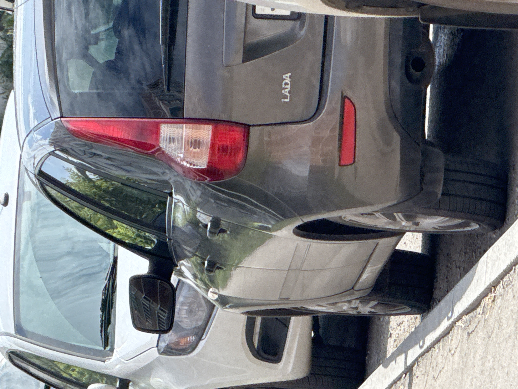 The image shows two cars parked on a street in Perm, Russia. The car in the foreground is dark gray and has "LADA" badging on the rear. The car behind it is white. Both cars are parked parallel to a concrete curb. The scene appears to be an outdoor urban environment on a sunny day.