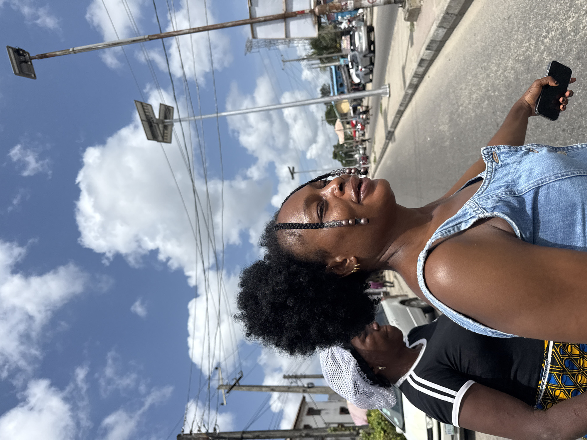 A medium shot captures a Black woman with her head tilted back, possibly taking a picture with her phone in her right hand. She has dark skin, dark curly hair pulled back into a bun with braids, and a denim top. Another person partially visible behind her is wearing a white cap, a black shirt, and a patterned yellow and blue cloth tied around the waist. The scene takes place on a city street in Ado, Nigeria. Visible in the background are power lines, streetlights, buildings, and parked cars, suggesting a bustling urban environment. The sky above is a mix of blue and white clouds.