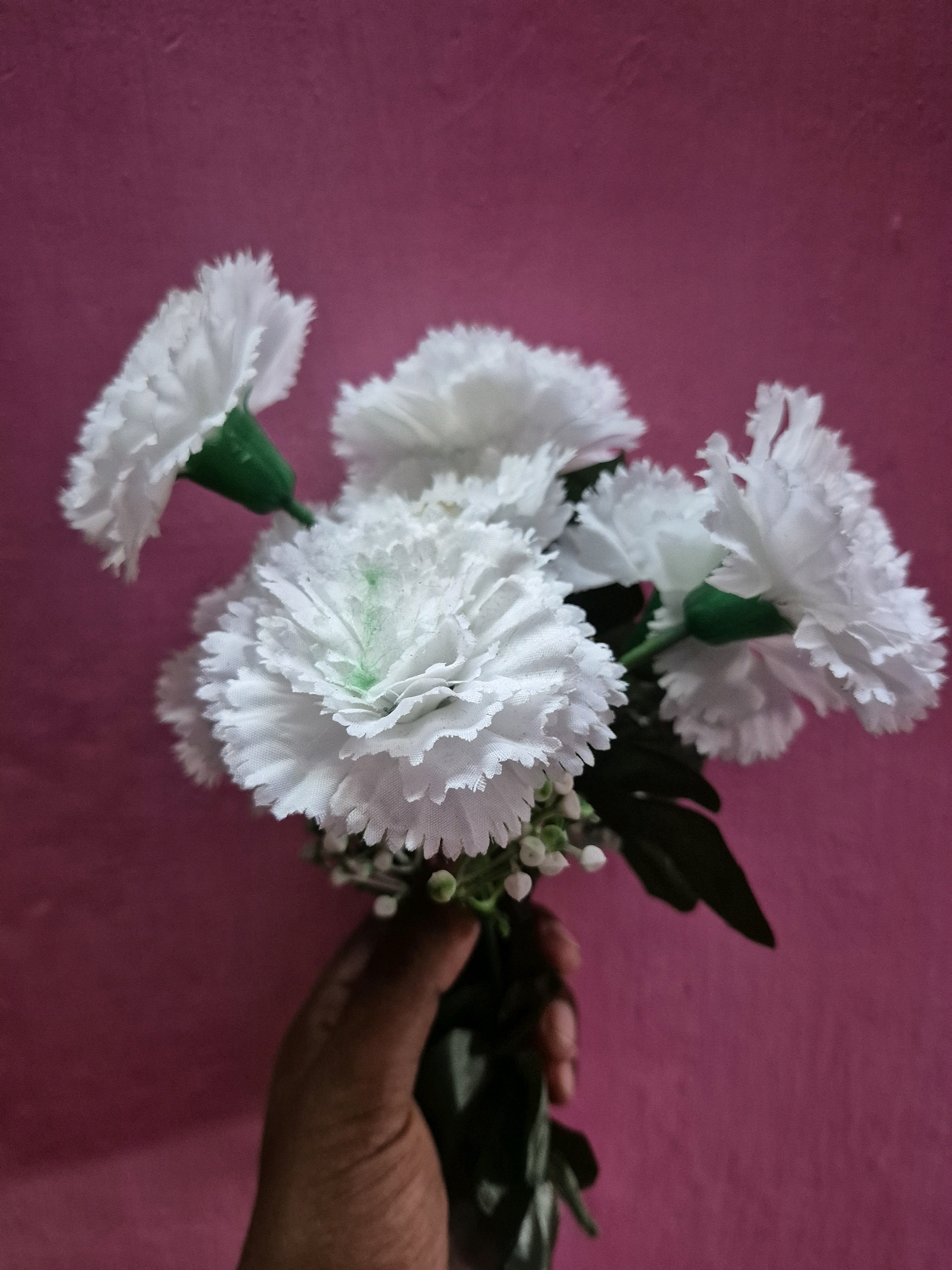 A hand is holding a bouquet of white artificial carnations against a pink textured wall. The flowers have green accents on their stems and some have a slight green tint in the center of the petals. The bouquet is arranged in a way that the flowers are clustered together and facing the viewer. The background is a solid pink wall with a subtle texture. There is no discernible information in the image that could indicate a location in Sambalpur, India.