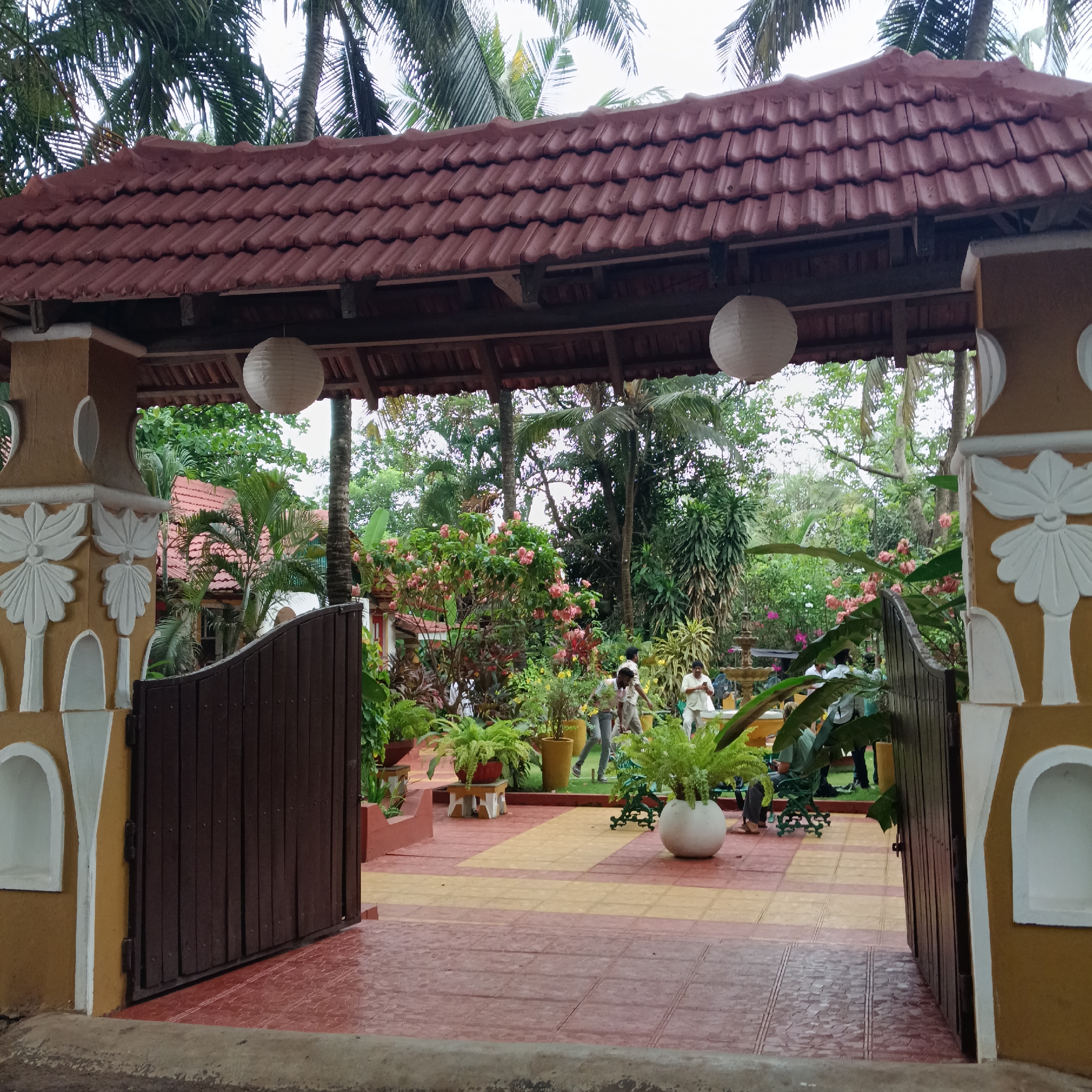 The image depicts the entrance to a property in Anjuna, India. The entrance features a covered gateway with a reddish-brown tiled roof, supported by yellow-painted columns adorned with white decorative elements. Two white paper lanterns hang from the structure. A wooden gate is partially open, revealing a courtyard paved with alternating red and yellow tiles. The courtyard is decorated with potted plants, including ferns. In the background, people are engaged in activities near a fountain amidst lush greenery and palm trees. A building with a reddish roof is also visible in the distance. The overall environment suggests a well-maintained garden or resort.