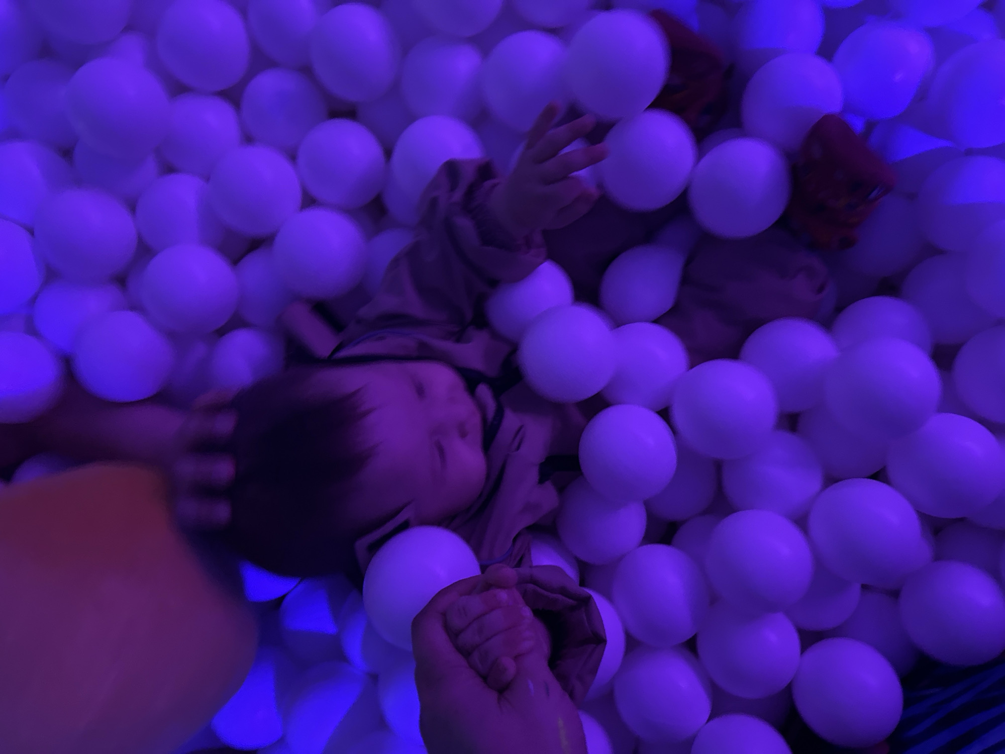 An infant or young toddler is situated in a ball pit, illuminated by prominent purple and blue artificial light. The setting is an indoor recreational area in Unknown, Unknown. The child is positioned centrally, lying or sitting amidst numerous white, spherical balls which appear purple due to the ambient lighting. They wear a light-colored long-sleeved garment and dark trousers. Dark hair covers their head, and their gaze is directed downwards and to the right. An adult's hand (light skin tone) on the left side of the frame holds the child's left hand. The child's right hand is extended forward with splayed fingers, appearing to interact with the surrounding balls. In the upper portion of the image, a segment of another adult's peach-toned arm or torso is visible, with fingers resting gently on or near the child's head or upper back. The child's feet, fitted with red shoes or socks, are visible towards the bottom right, partially submerged among the balls. The scene depicts a supervised interaction between the child and two adults within the brightly illuminated ball pit environment.