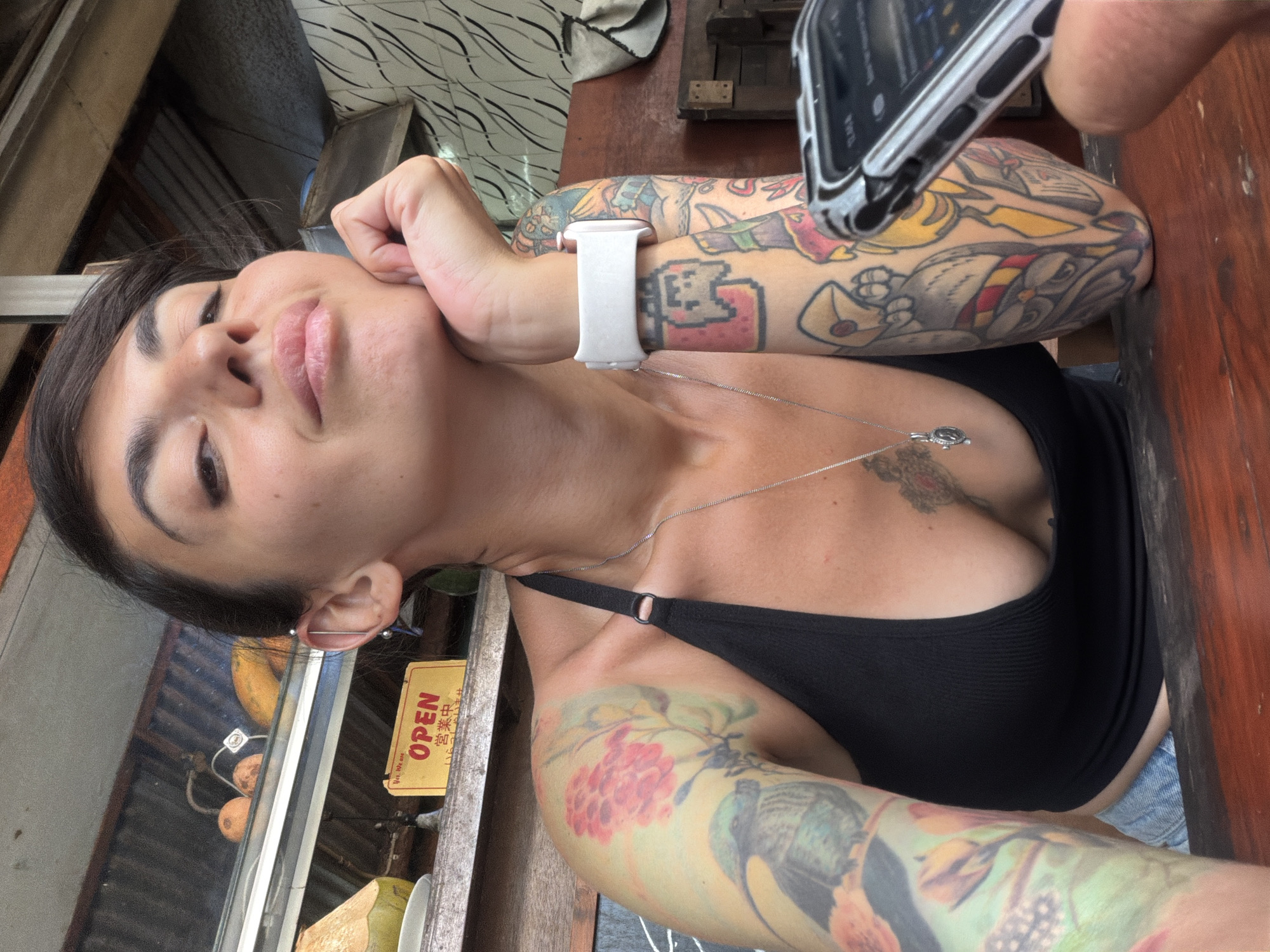 A woman with dark hair, a black tank top, and numerous colorful tattoos is posing for a selfie at a wooden table. She is leaning on her hand, looking towards the camera with a relaxed expression. In the background, a yellow sign with red lettering that says "OPEN" is visible, along with some fruits and a coconut. The scene appears to be outdoors, possibly at a food stall or market in Kuta Selatan, Indonesia.