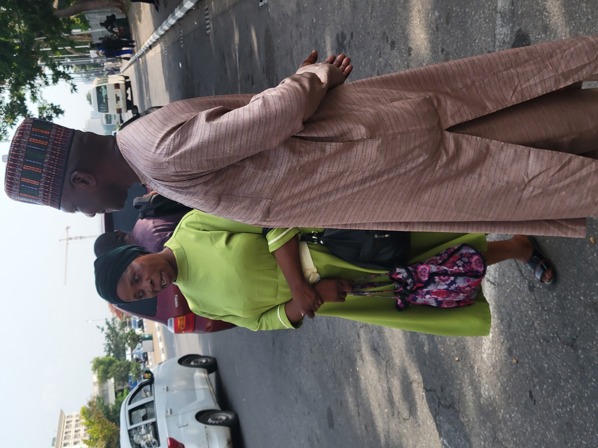 The image depicts two individuals standing on a city street in Abuja, Nigeria. The taller individual is a man wearing a brown, striped, long-sleeved garment and a traditional Nigerian hat. He is looking down. The other person is a woman wearing a green dress, a black head covering, and sandals. She is carrying a black handbag and a flowered bag.
They appear to be near a road with moving traffic, including white and red cars. A building is visible in the background, along with a construction crane. The street is paved with visible shadows, suggesting it is daytime.