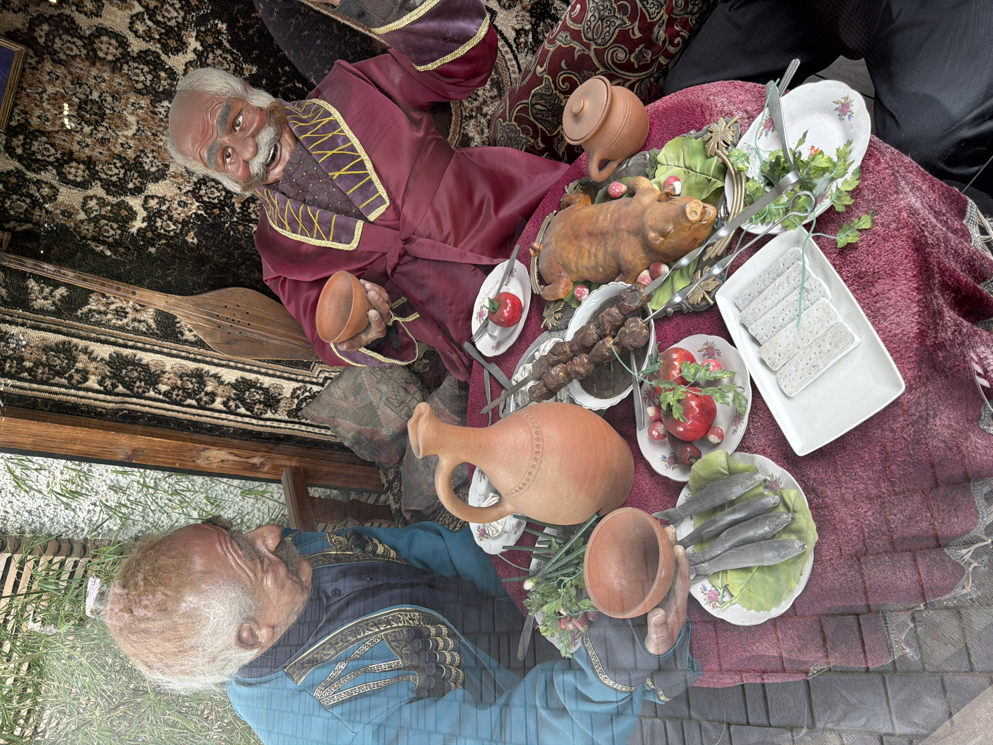 The image captures a still life display, likely within a shop window or museum exhibit, in Tbilisi, Georgia. Two lifelike mannequins, appearing as elderly men, are positioned as if sharing a meal. The mannequin on the left wears a teal, embroidered garment and is looking towards the right. The mannequin on the right is dressed in a maroon robe with gold trim and is holding a small, empty clay bowl, with his mouth open in a jovial expression.
In front of the mannequins is a table covered with a red, textured tablecloth, laden with food items. Several dishes are present: a platter of four whole fish on lettuce leaves, a smaller plate with two red tomatoes and some greenery, a platter featuring a whole roasted piglet surrounded by small, red mushrooms, and a long skewer of grilled meat. Also on the table are several pieces of white, rectangular food items, possibly bread or cheese, arranged on a white rectangular platter. A large, terracotta jug and a smaller terracotta pot are also visible.
The background depicts an interior setting with woven rugs, wooden beams, and decorative elements, suggesting a traditional or cultural context. The presence of a stringed instrument, possibly a lute or a saz, leaning against the wall further reinforces this impression. The overall scene is static, depicting a posed arrangement rather than a candid moment. The lighting suggests an indoor environment, possibly with natural light filtering in from the left.