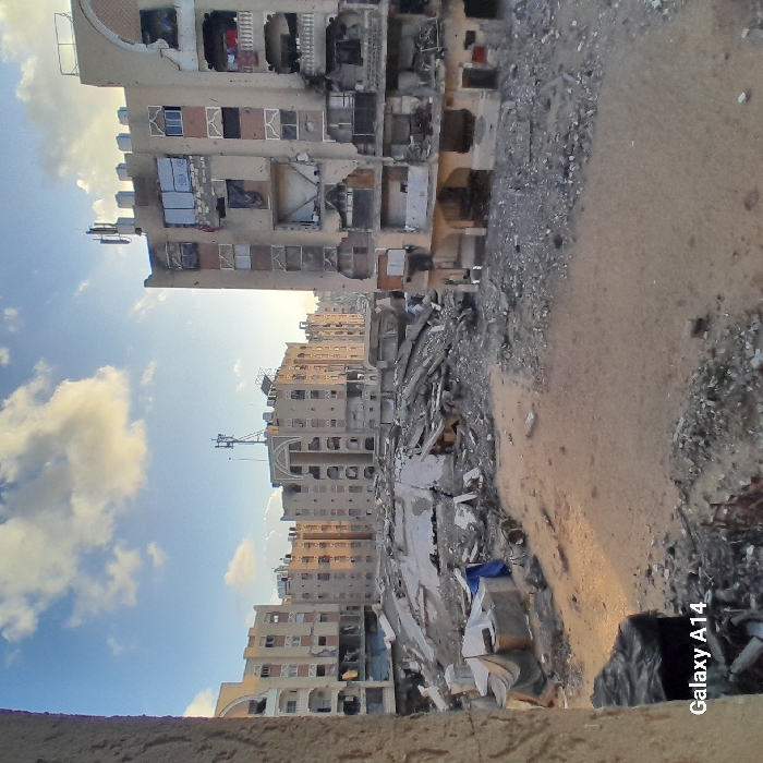 The image shows a devastated cityscape. Buildings are severely damaged, with many floors collapsed or riddled with holes. Rubble and debris cover the ground. A mix of tan and grey dominates the color palette, suggesting concrete and dust. The sky is visible with clouds, and one building is heavily damaged. The Samsung Galaxy A14 watermark is visible in the lower right corner.