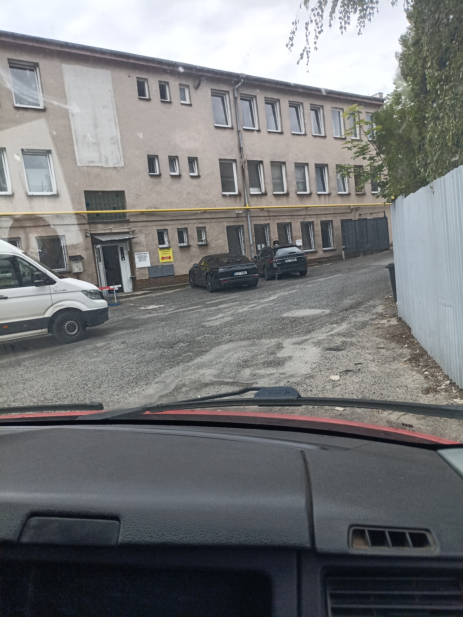 The image shows a view from inside a vehicle looking out at a building and parked cars in what appears to be an industrial or commercial area in Svémyslice, Czechia. A light-colored van is parked on the left, and two dark-colored cars are parked further down on the right. The building behind them is a multi-story structure with numerous windows. The ground is paved with a rough, textured surface, and there is a tall, corrugated metal fence on the right side of the frame. The sky is overcast. The interior of the vehicle, including the dashboard and windshield wipers, is visible in the foreground.