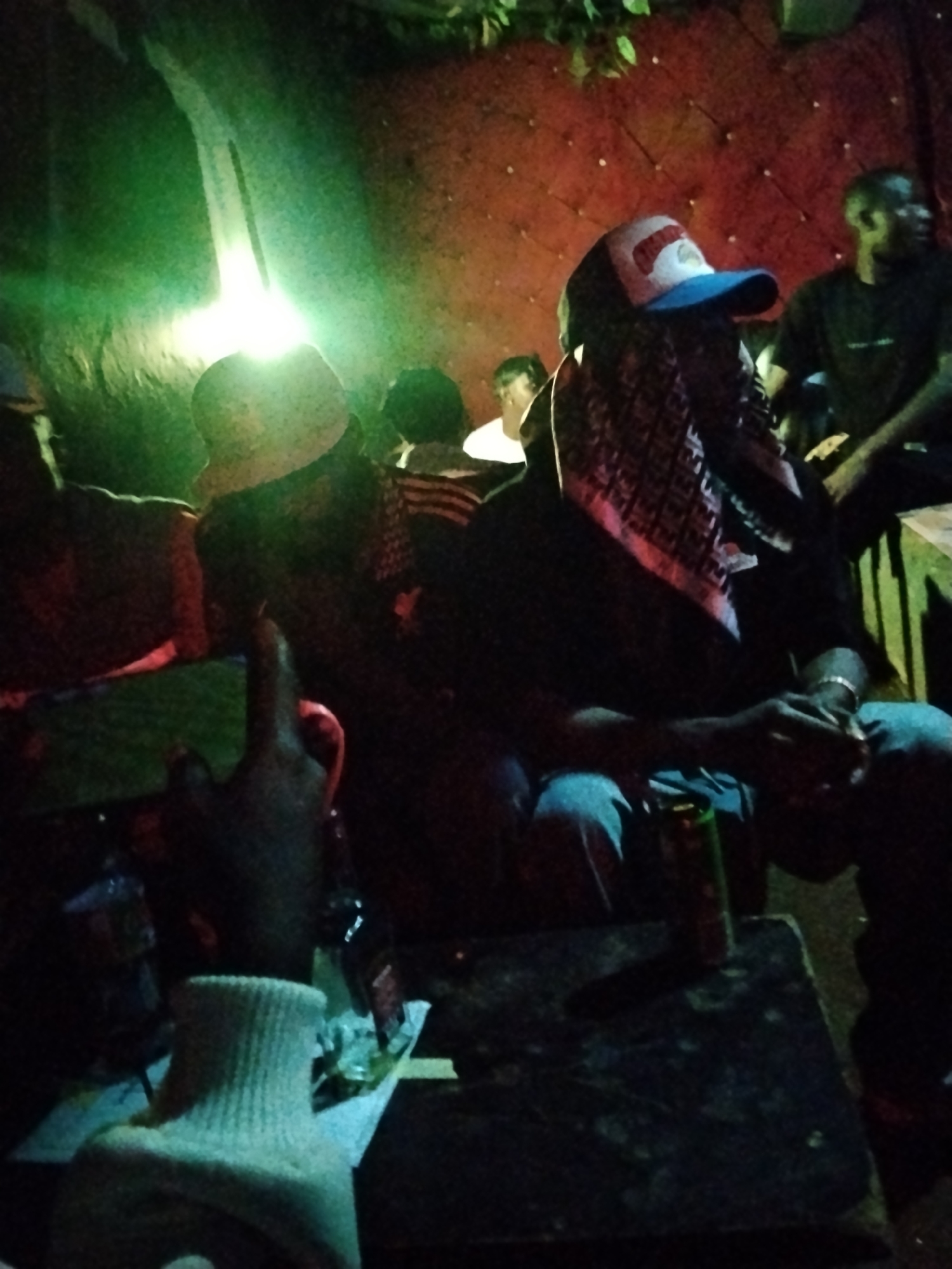The image shows a dimly lit indoor scene, likely a social gathering. In the foreground, a person wearing a white ribbed turtleneck and a black sleeve is partially visible, with their hand raised and pointing. They are positioned to the left of a dark, textured surface, possibly a table, on which a green and silver can is placed. Further back, a person is seated, wearing a blue and red baseball cap and a patterned headscarf or cloth draped over their head and shoulders. This individual is also wearing a dark long-sleeved shirt and blue jeans. Their hands are clasped in their lap, and a silver bracelet is visible on their right wrist. To their right, another person is seen wearing a black t-shirt. The background features a textured red wall with circular indentations, and hints of green foliage are visible at the top left, suggesting an outdoor or semi-outdoor element. The lighting is uneven, with a bright green light source on the left casting shadows and highlighting certain areas. The overall atmosphere appears casual and relaxed. The image context suggests this scene is located in the city of Durumi, Nigeria, though no specific landmarks or contextual clues within the image itself confirm this location.