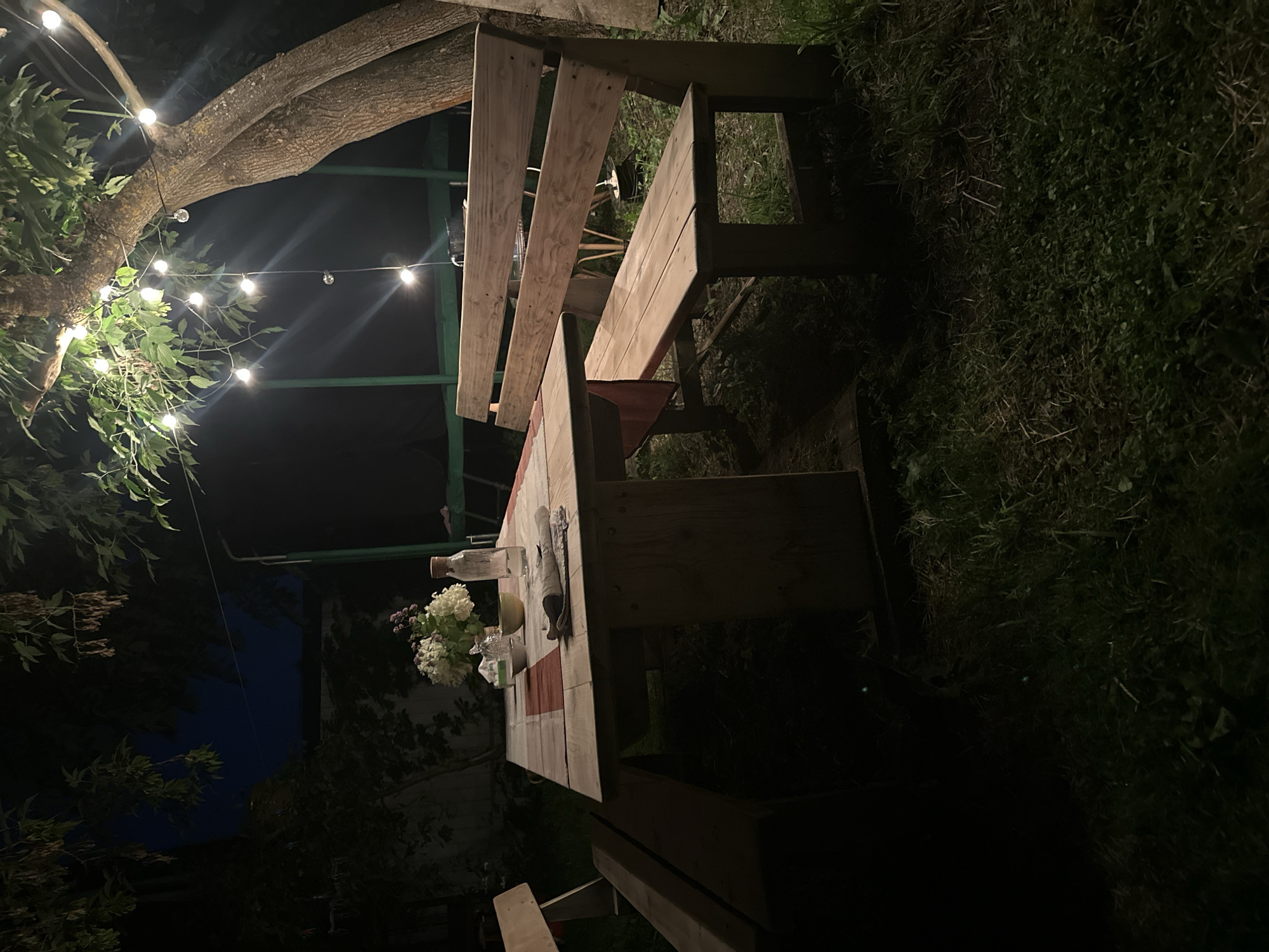 This is a nighttime outdoor scene, likely in a backyard in Staritsa, Russia, as indicated by the presence of a rustic wooden picnic table with benches, a bouquet of white flowers in a vase, a bottle of water, and a folded napkin on the table. String lights are visible overhead, illuminating the scene from a large tree. The ground is covered in grass, and the sky is dark. There are no people present. The overall impression is one of a peaceful, unlit evening gathering space.