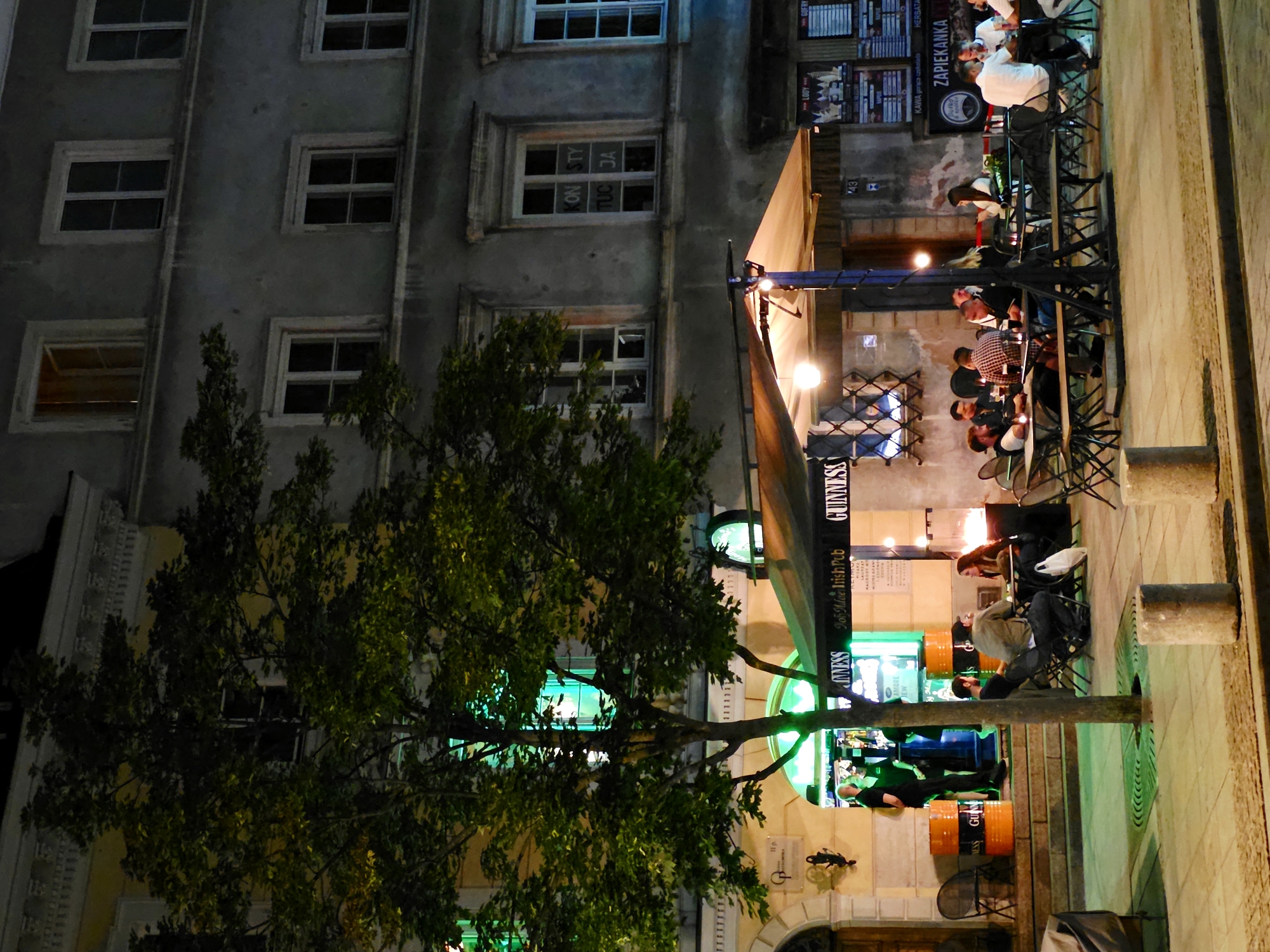 The image captures a nighttime scene outside a pub in Warsaw, Poland. The pub, identified by signage for "GUINNESS" and "Irish Pub," appears to be located on a pedestrianized street with cobblestone paving. A prominent tree stands in the foreground, its branches casting shadows across the sidewalk and the outdoor seating area.
Several people are seated at tables, engaged in conversation and enjoying drinks. Some tables are covered by an awning, illuminated by overhead lights, while others are exposed to the ambient street lighting. The building facade behind the pub features multiple windows, some of which are lit from within, indicating residential or commercial spaces above. The overall atmosphere suggests a lively evening at a popular establishment.