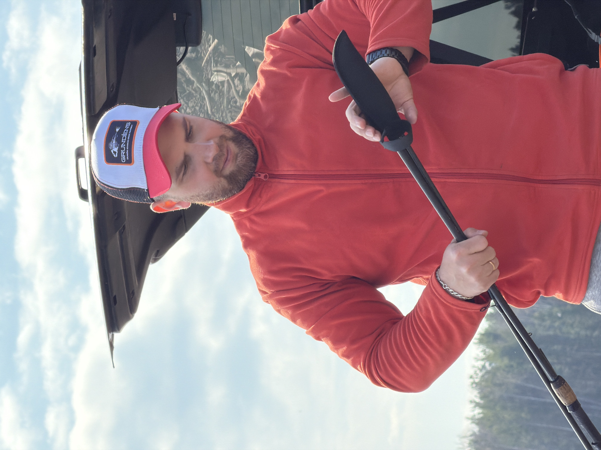 A man is seen in an outdoor setting, likely near a body of water. He is wearing a red zip-up jacket and a white and red baseball cap with a logo on the front. He holds a black object in his right hand and a fishing rod in his left. The backdrop features a vehicle with an open trunk and a landscape of trees. The location details are absent from the image.