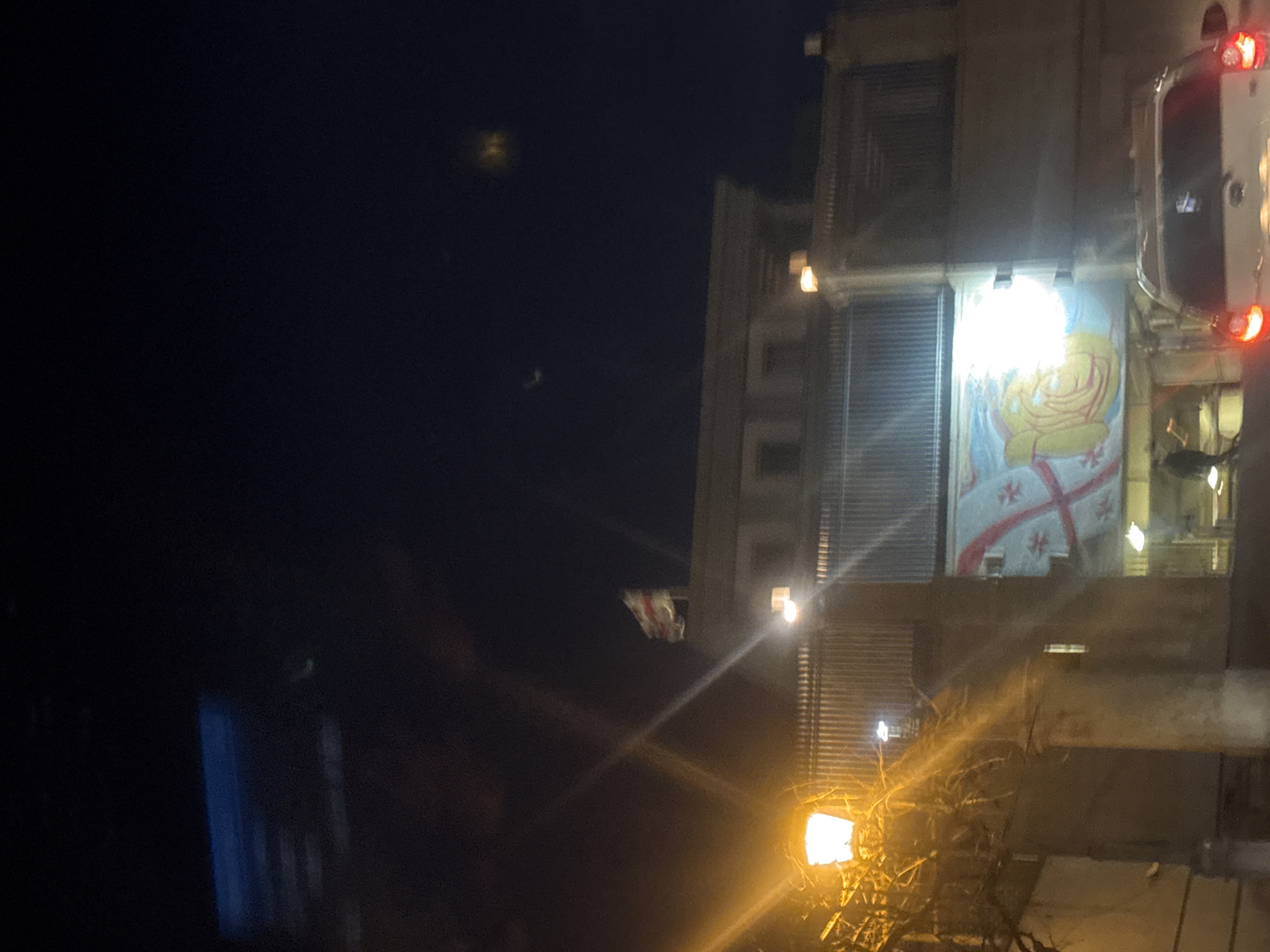 At night, the image depicts a building facade in Tbilisi, Georgia. A flag, possibly Georgian, is displayed on the building. An illuminated banner with a religious motif is visible. A street lamp provides lighting. A vehicle with illuminated tail lights is partially visible on the right.