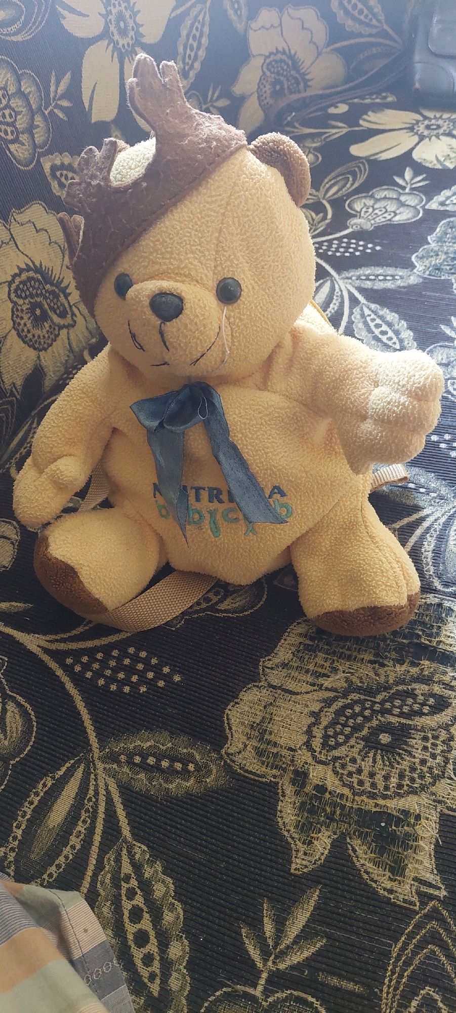 A light yellow teddy bear wearing a brown antler headband is sitting on a dark patterned couch. The bear has black button eyes and a black nose. A thin white line runs from under its right eye to its mouth, resembling a tear. A dark blue ribbon is tied in a bow around the bear's neck. The bear has "MATRIA BJYC" embroidered in blue thread on its chest. A light beige strap is visible across its back. The couch fabric has a dark background with a large, intricate floral pattern in yellow and lighter shades. A folded striped blanket is partially visible in the bottom left corner of the image. This scene is likely indoors, possibly in a residential setting within Lumajang, Indonesia.