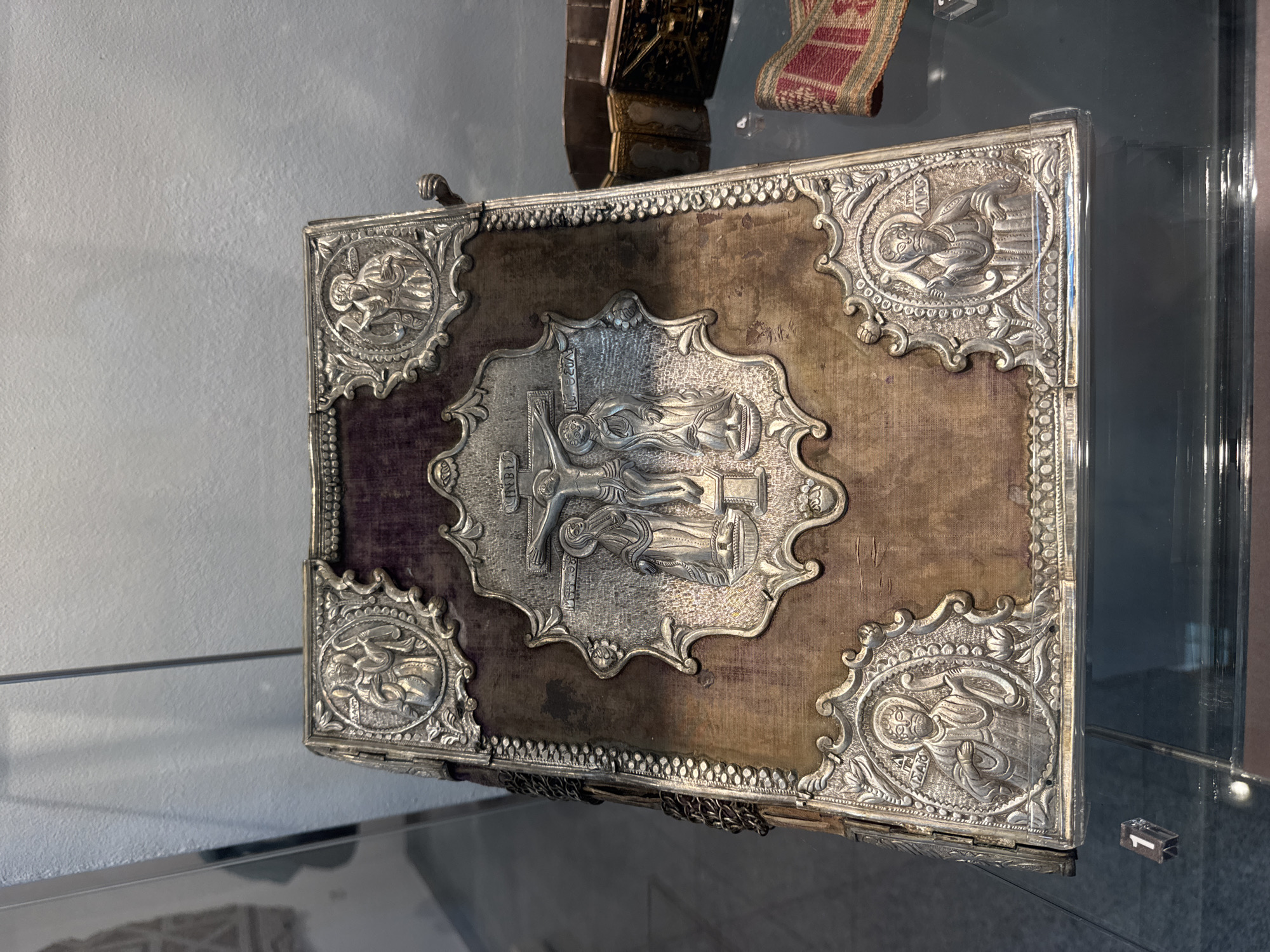 The image shows a richly ornamented book in a glass display case, likely in a museum. The book's cover is adorned with silver detailing, including corner embellishments and a central depiction of the crucifixion. The background surface of the cover appears to be a dark velvet or similar fabric. The scene is located in the city of Plovdiv, Bulgaria. The environment is a museum setting, indicated by the display case. There are no people directly visible in the scene.