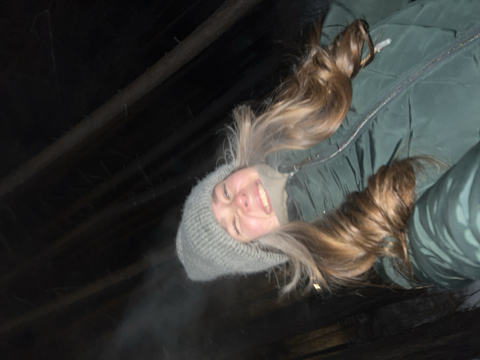 An individual is depicted in a close-up, low-angle selfie-style photograph taken at night in a snowy, forested area consistent with a nature park or reserve near Krasnoyarsk, Russia. The subject, a young adult with long brown hair, is smiling broadly, causing their eyes to squint. They are wearing a light grey knitted beanie, a dark green puffer jacket with a visible zipper, and a light beige neck gaiter. A wisp of visible breath or vapor is present near the subject's left cheek, indicating cold temperatures. The background consists of dark, vertical tree trunks, and a small patch of snow is visible at the bottom left of the frame. Illumination appears to be from a direct flash.